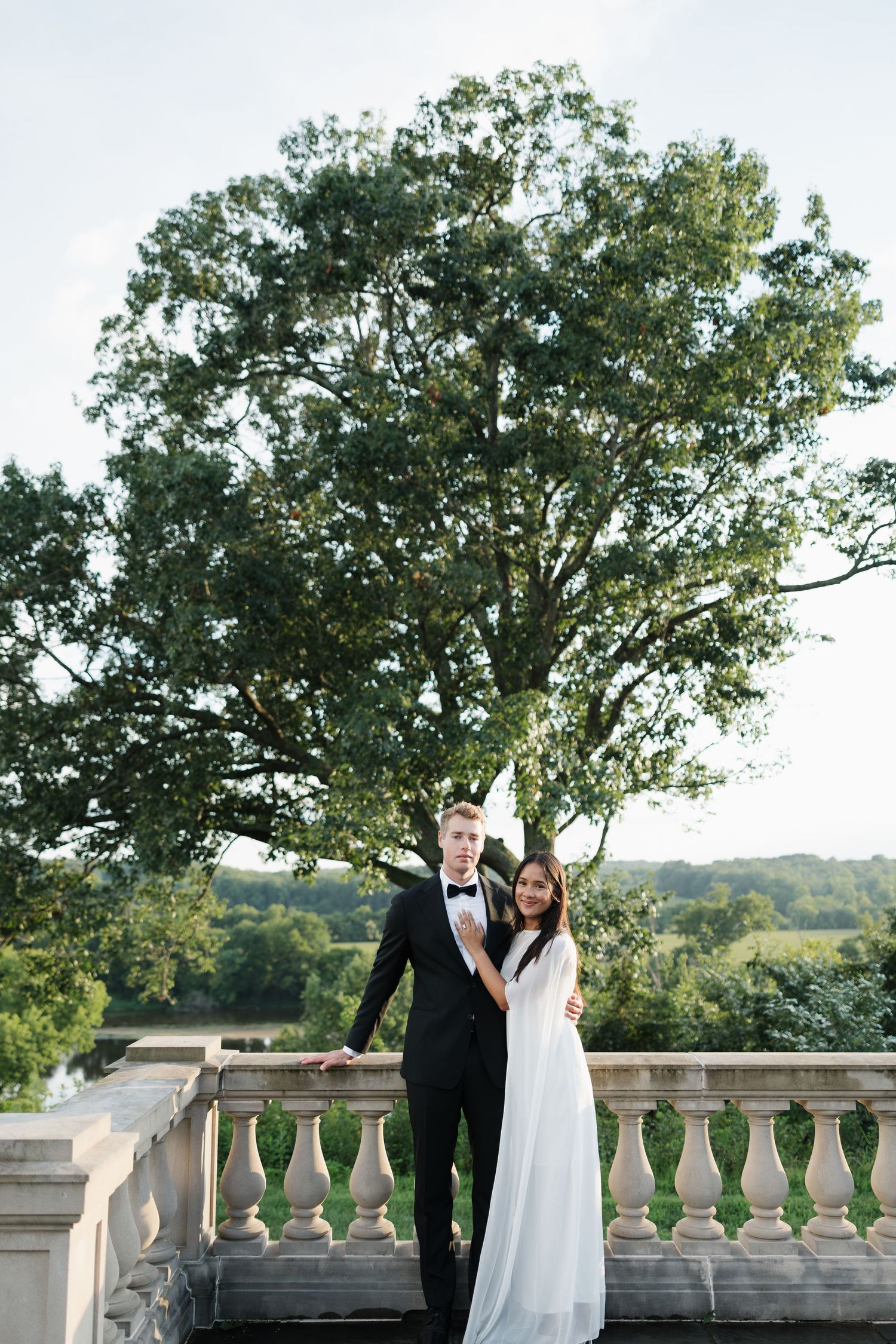 Bride and groom posing on a stone balcony with a large green tree and scenic countryside backdrop.