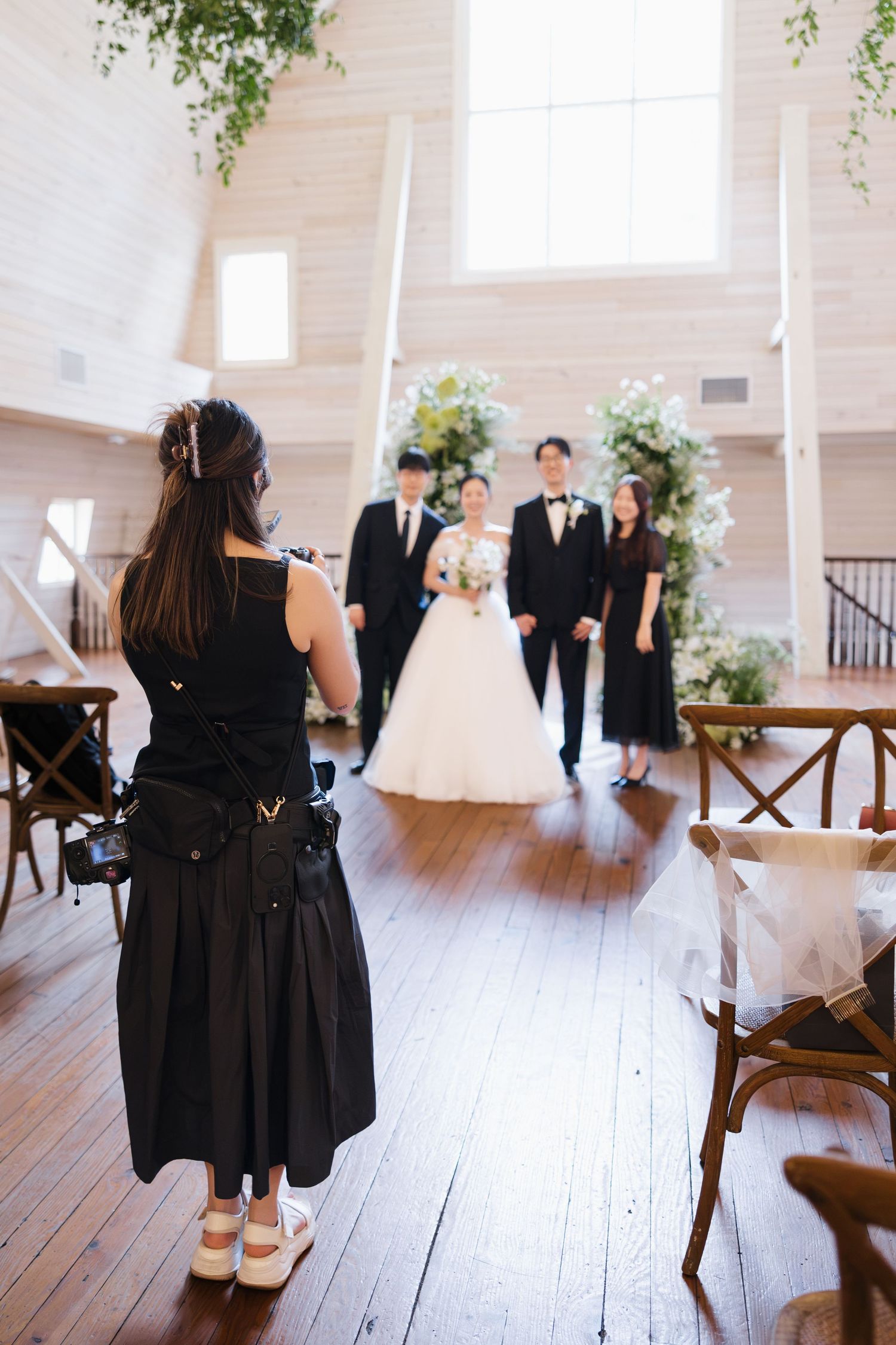 Photographer capturing bride and groom with guests in elegant white chapel with floral arch and hardwood floors.