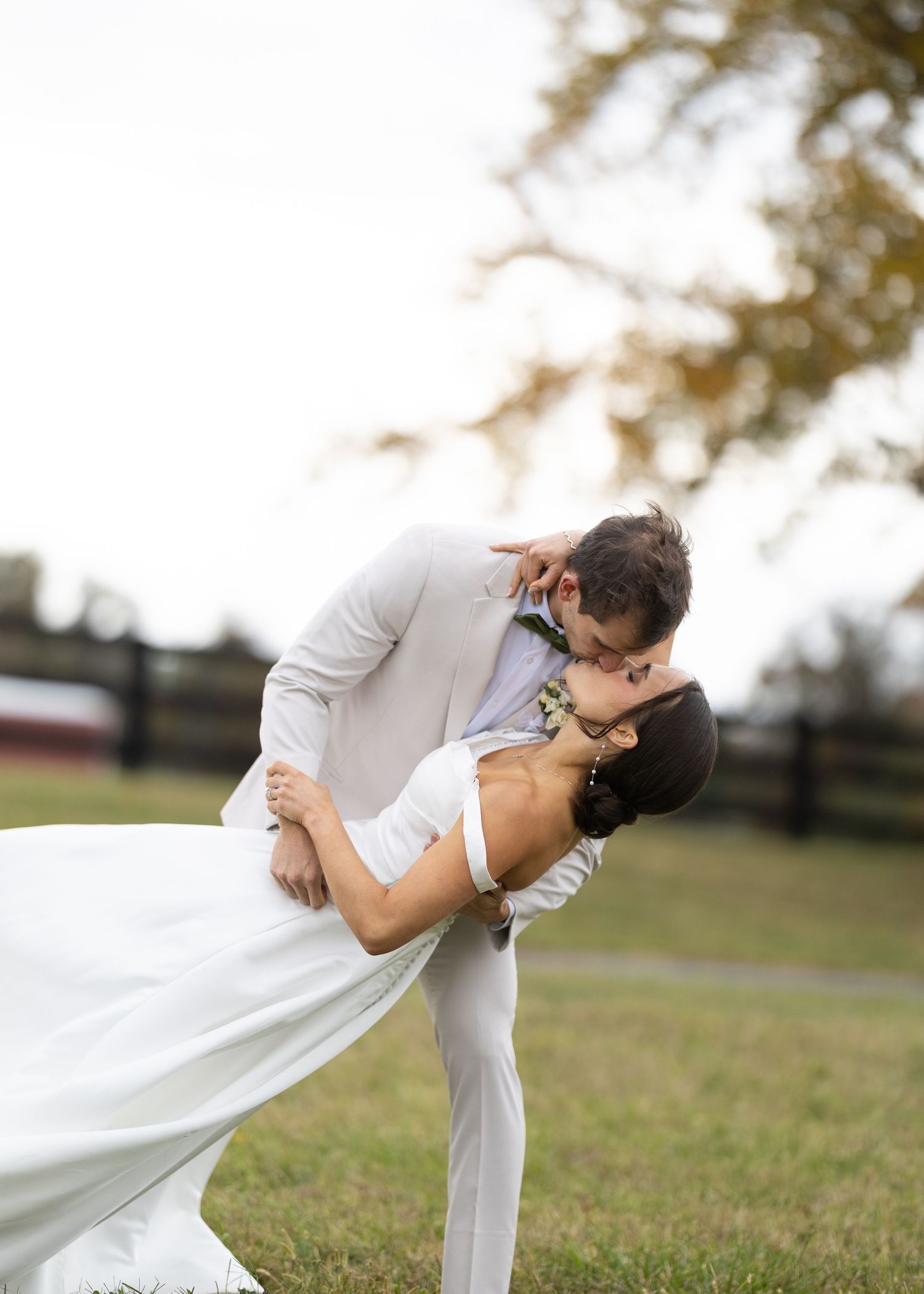 Groom dips bride for a romantic kiss outdoors on a green lawn with autumn trees in the background.