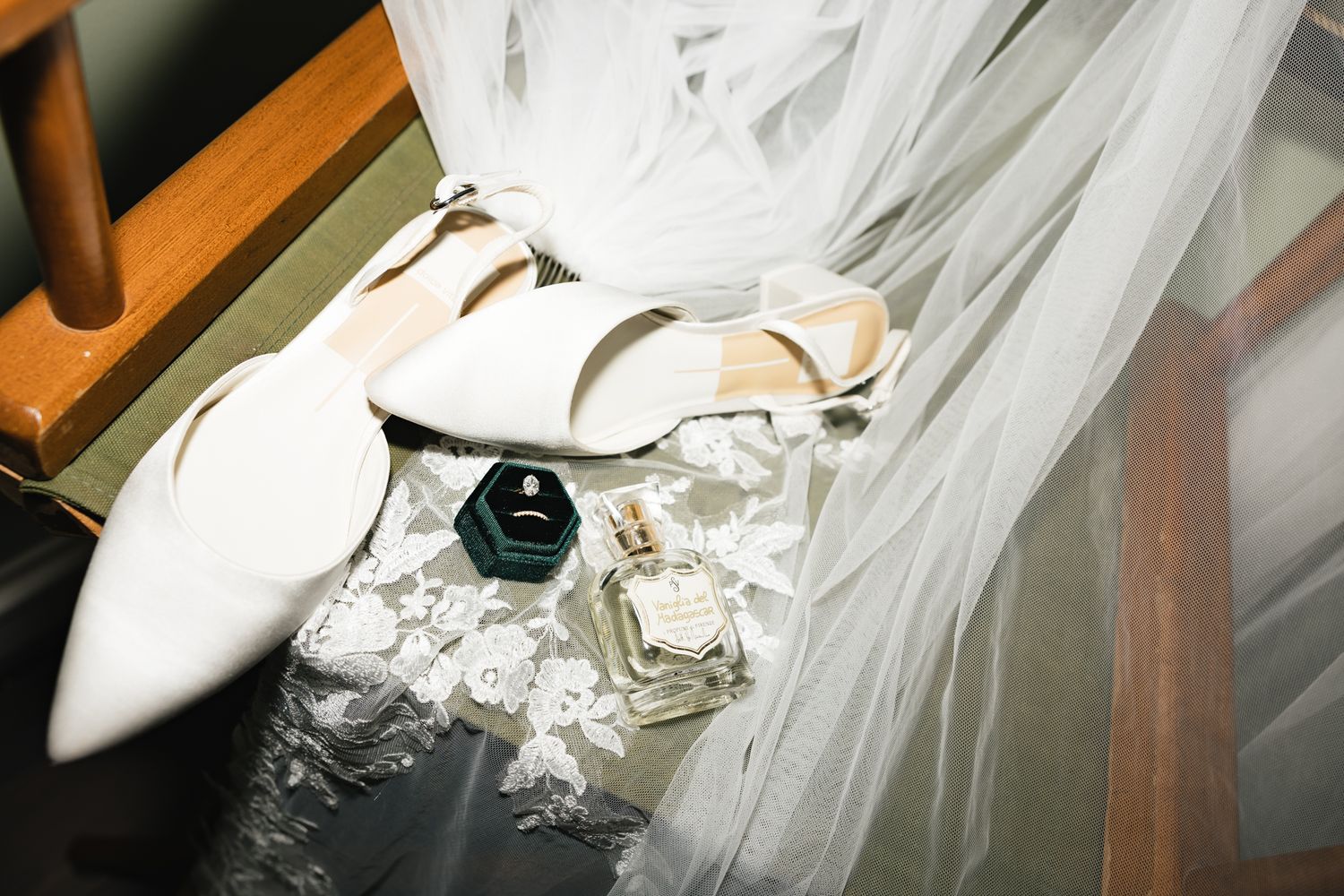 Elegant bridal accessories flat lay featuring white heels, engagement ring, and perfume bottle on lace fabric.