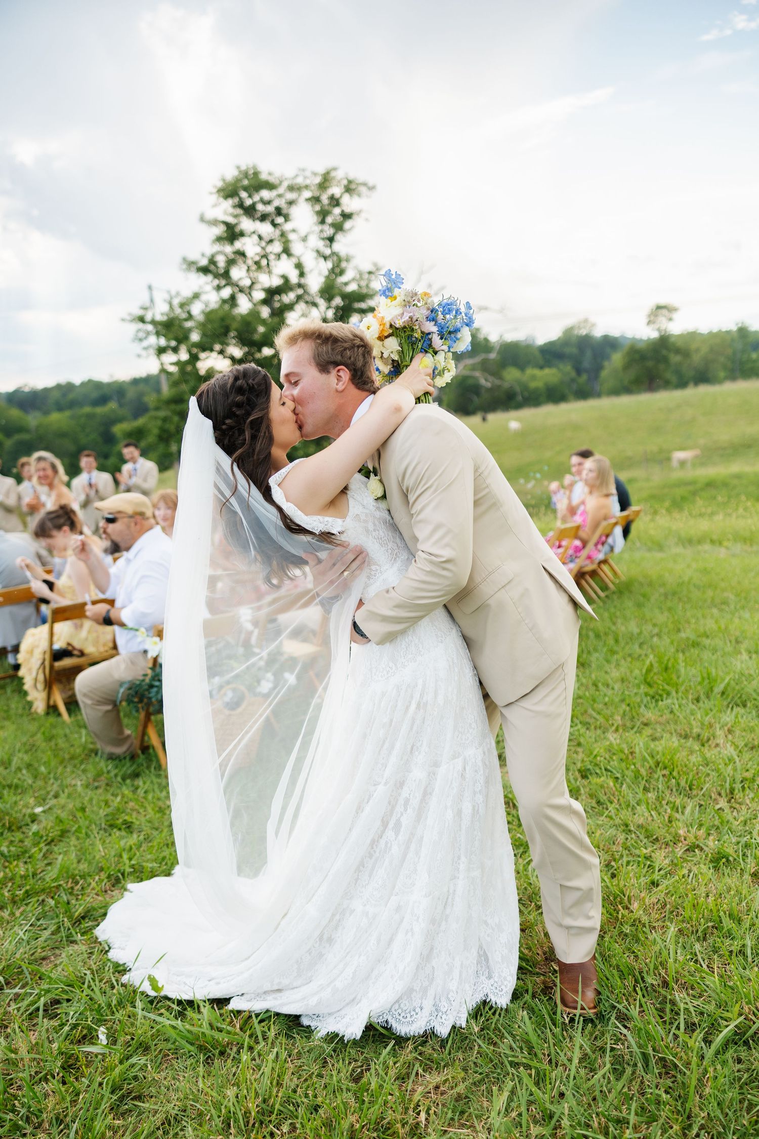 Bride and groom share first kiss at outdoor wedding ceremony on green grass field, bride holding colorful bouquet.