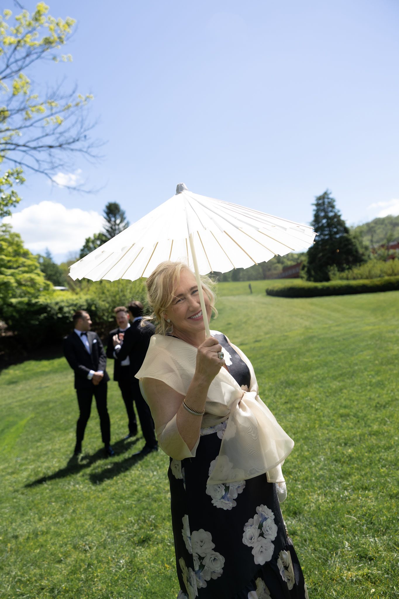 Woman in floral dress holding white parasol smiling outdoors at wedding reception on sunny day with green lawn.