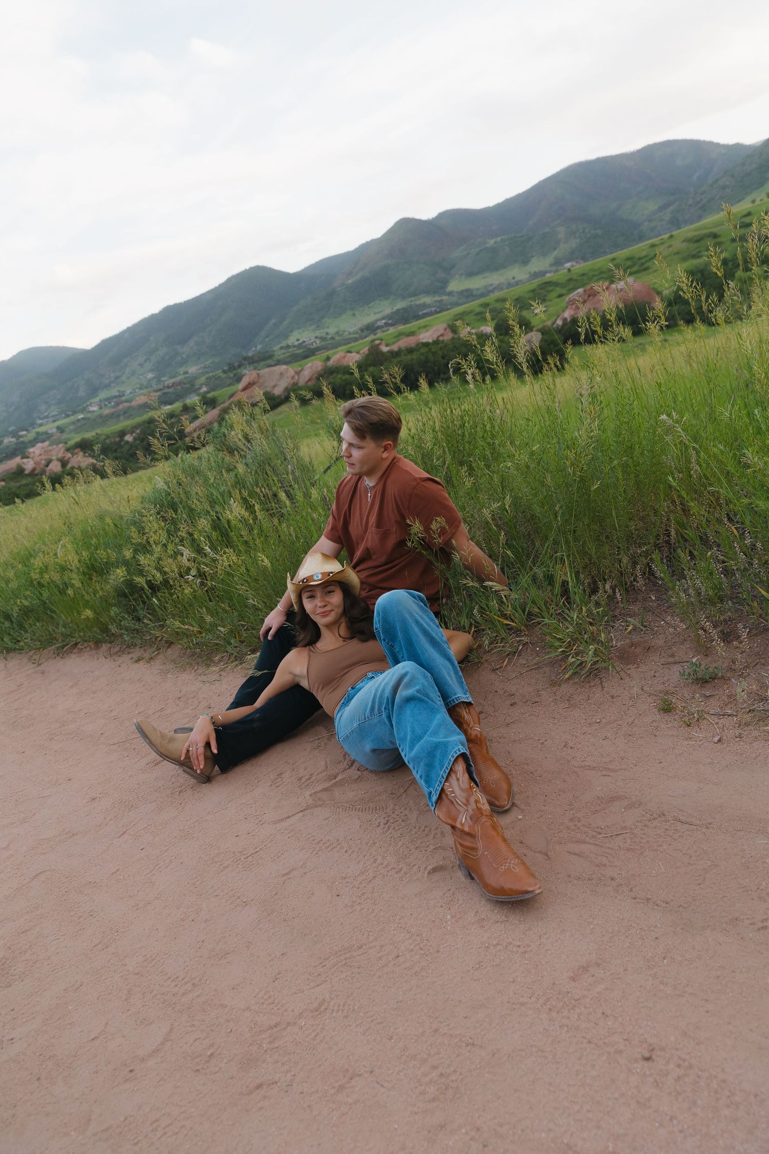 Couple relaxing on sandy dirt path with lush green mountain landscape in background.