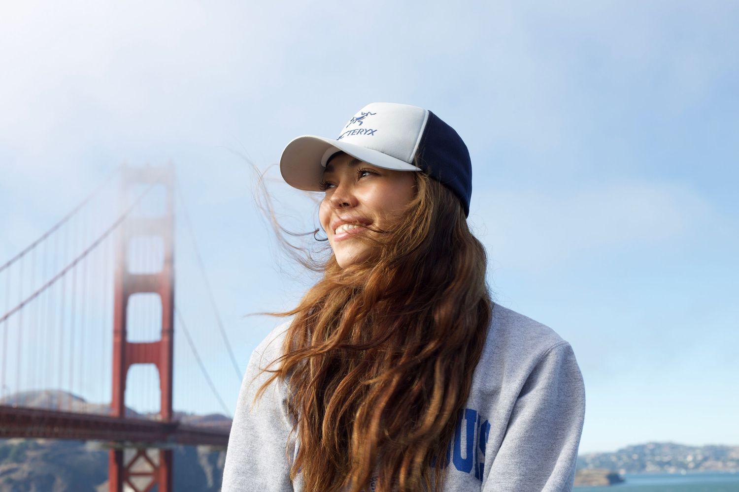 Young woman in baseball cap and hoodie with long brown hair near iconic Golden Gate Bridge on windy day.