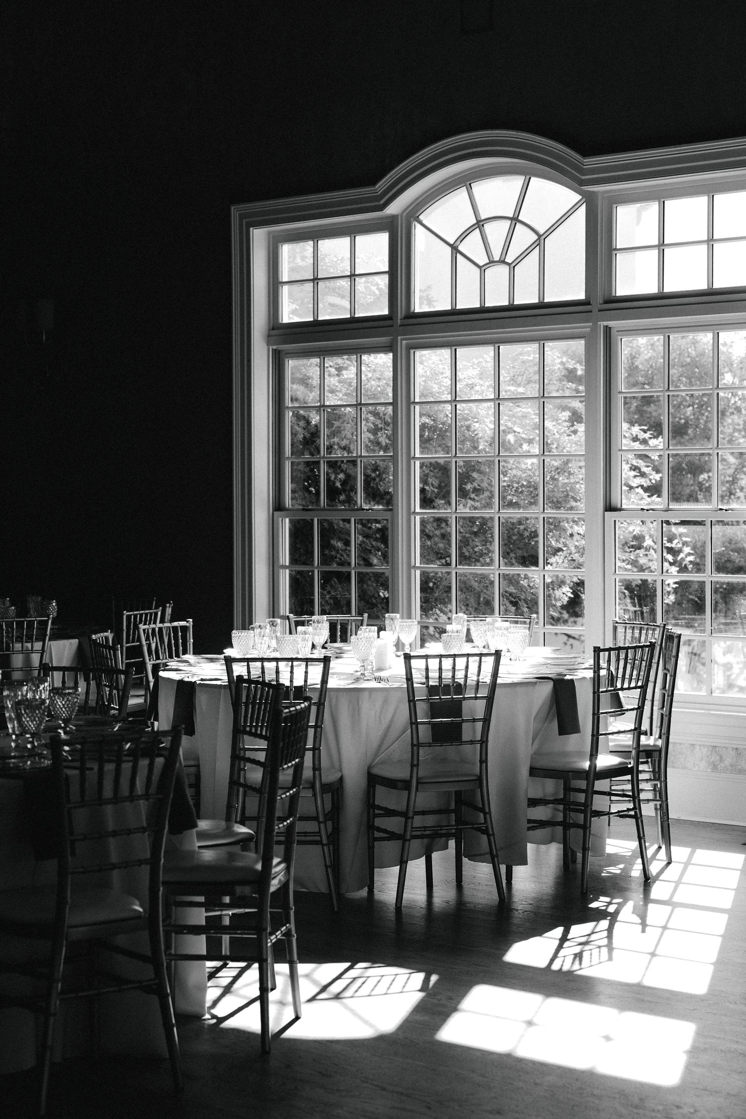 Black and white photo of an elegant banquet hall with round tables, chiavari chairs, and large arched windows overlooking greenery.