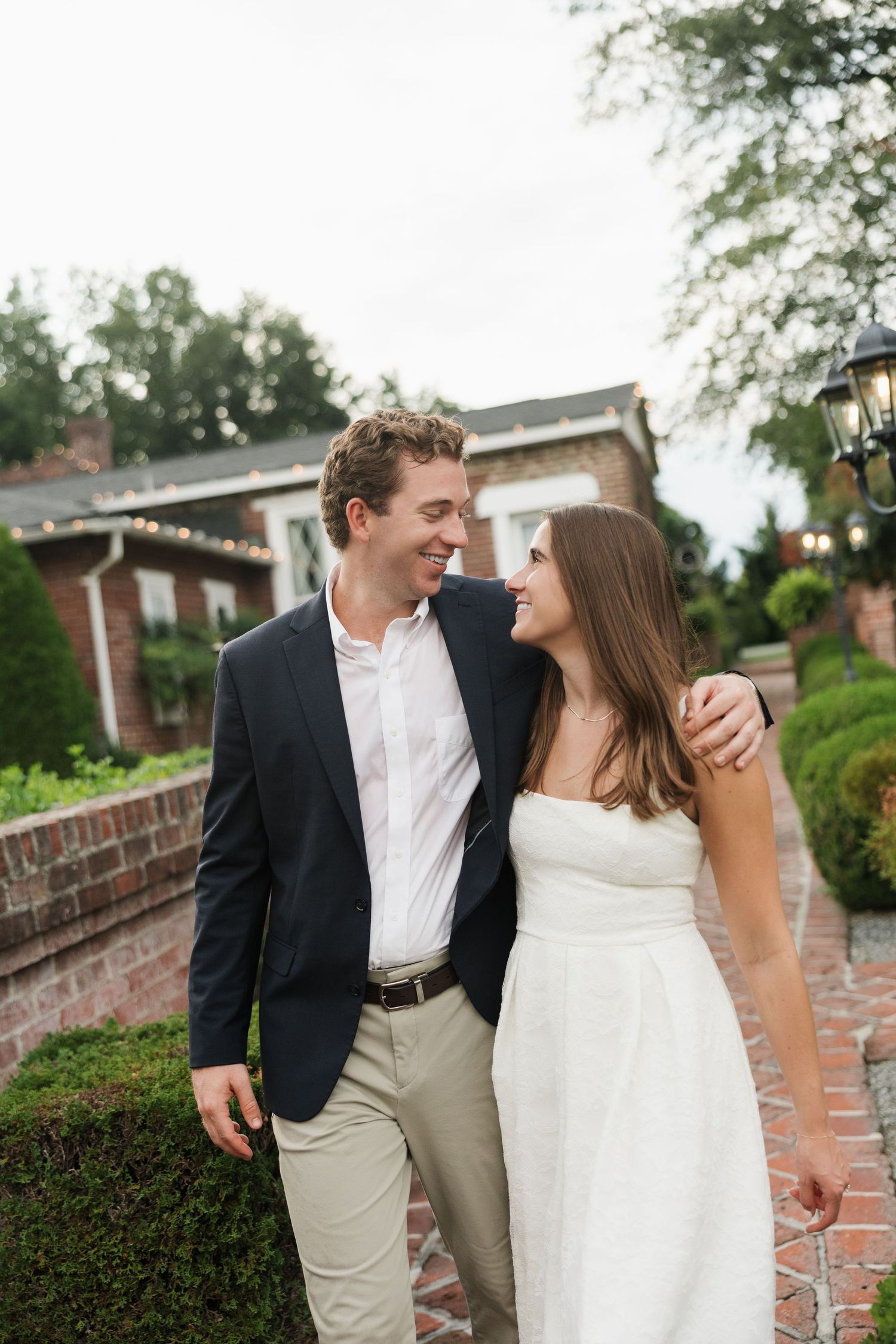 Smiling couple in elegant attire walking together outdoors near a brick building with lush greenery.