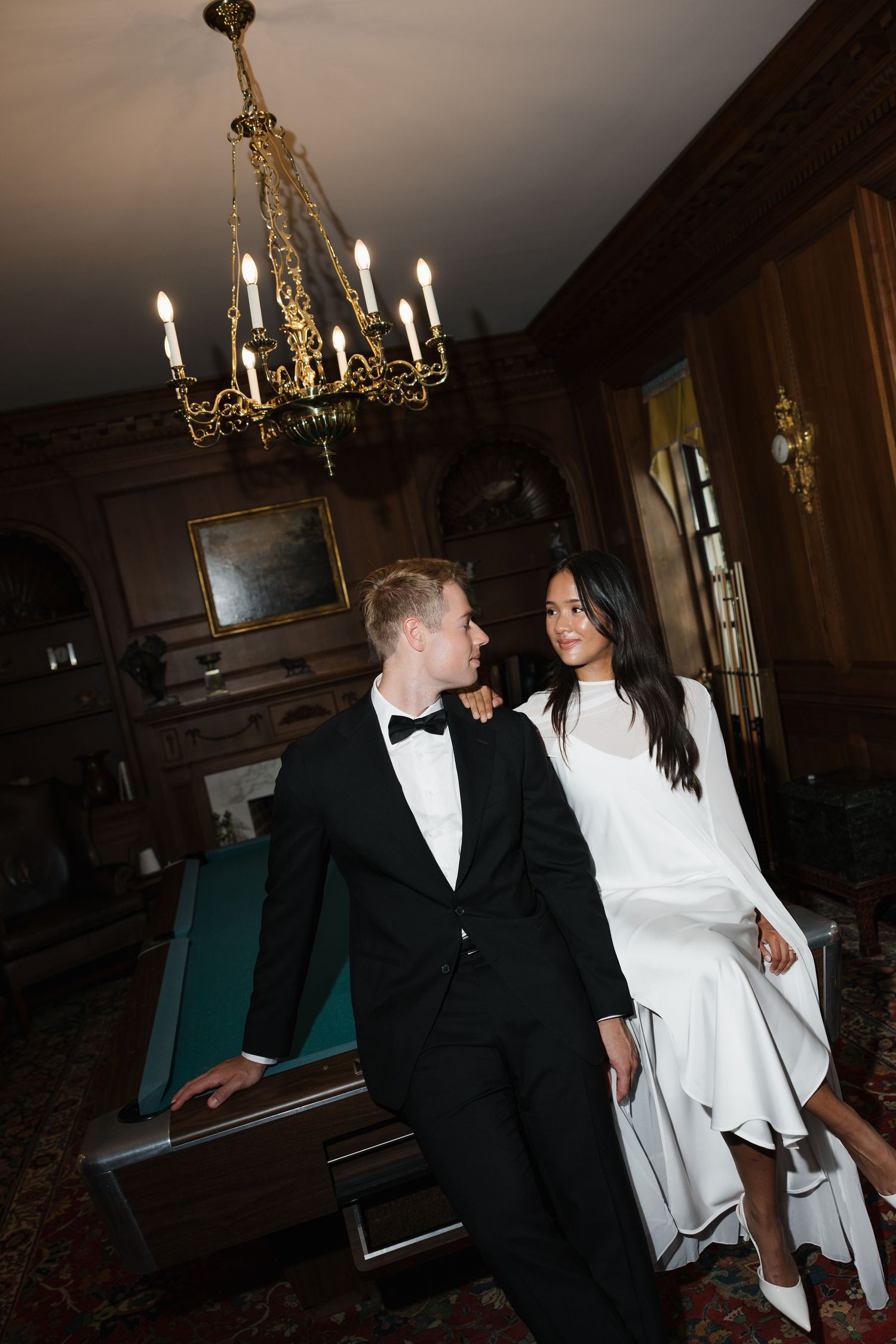 Elegant couple in formal attire, man in tuxedo and woman in white dress, posing in ornate wood-paneled room with chandelier.