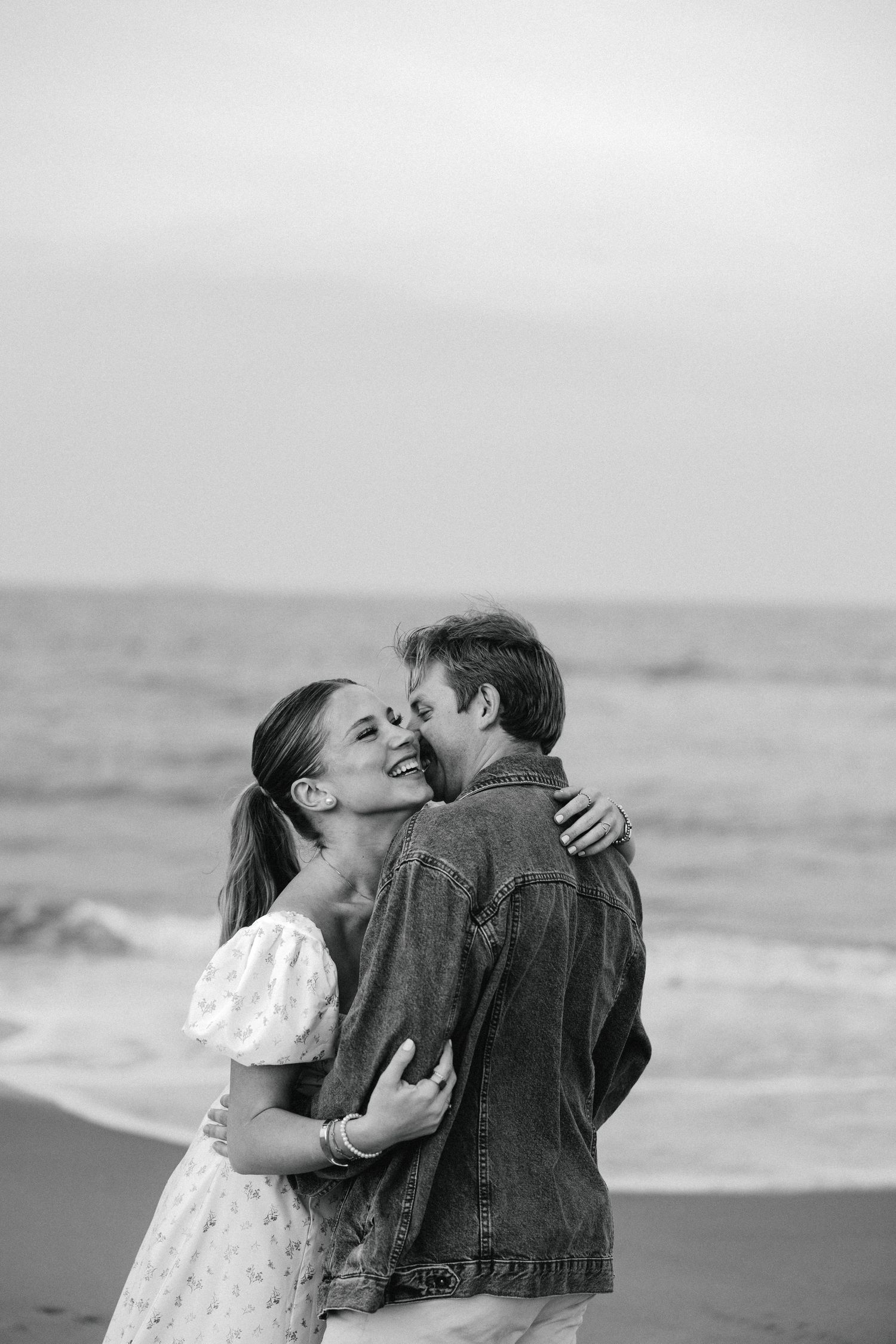 Black and white photo of couple hugging and laughing on beach with ocean in background.