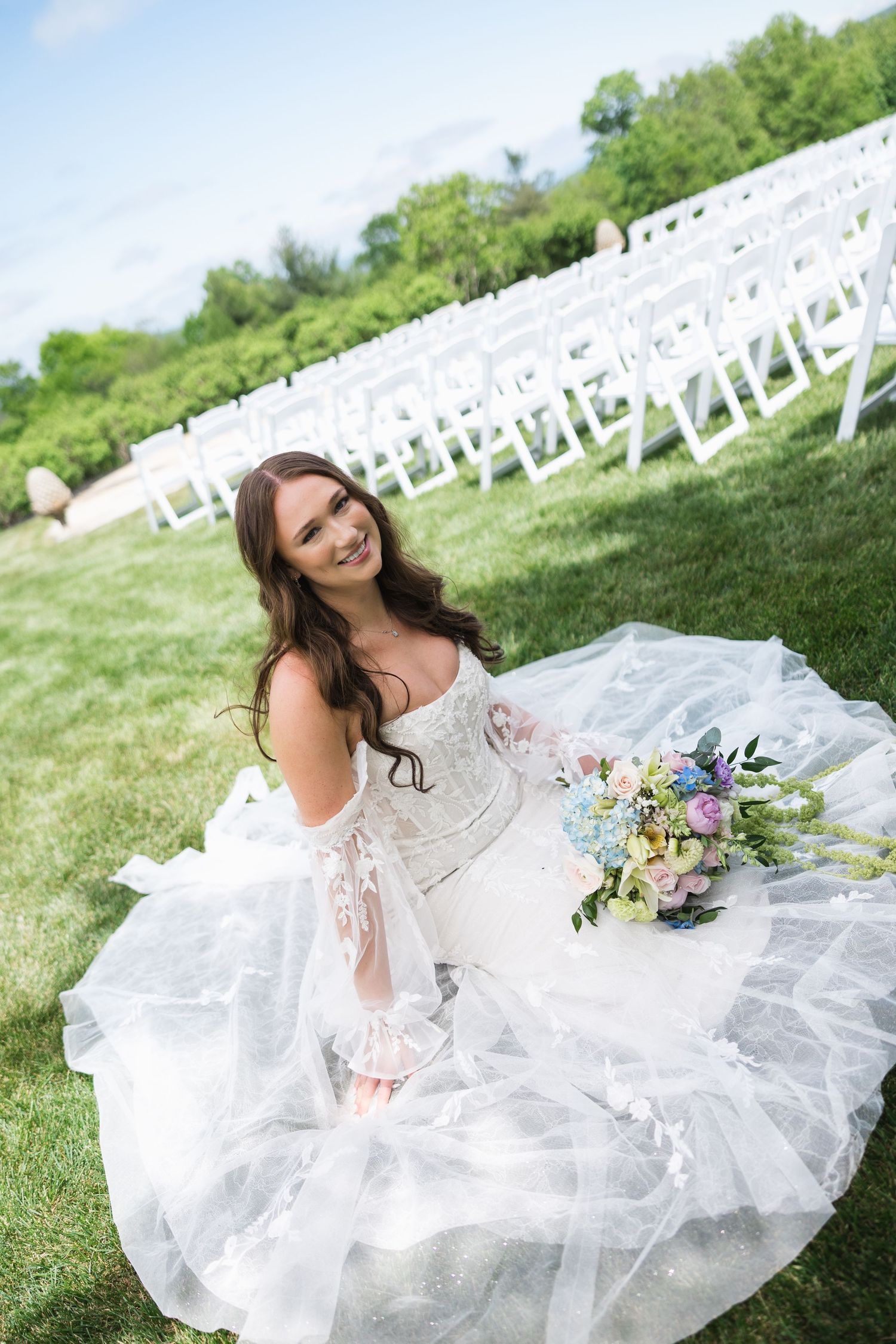 Bride in white lace gown sitting on grass with bouquet and white chairs.