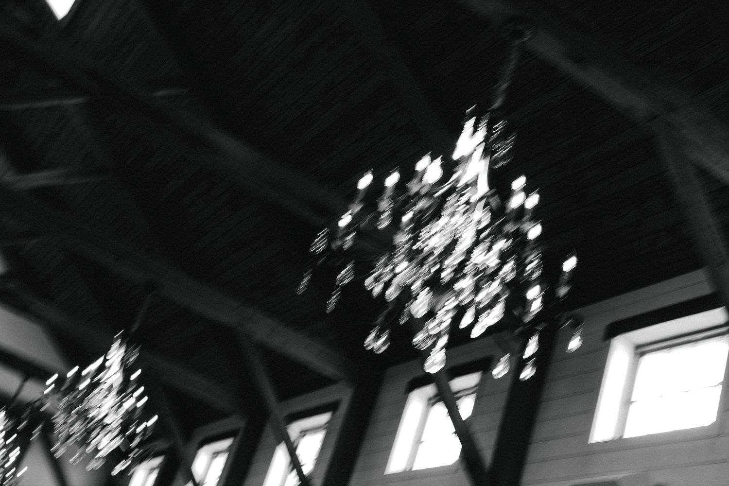 Black and white photo of decorative chandelier hanging from rustic wooden ceiling beams in wedding venue.