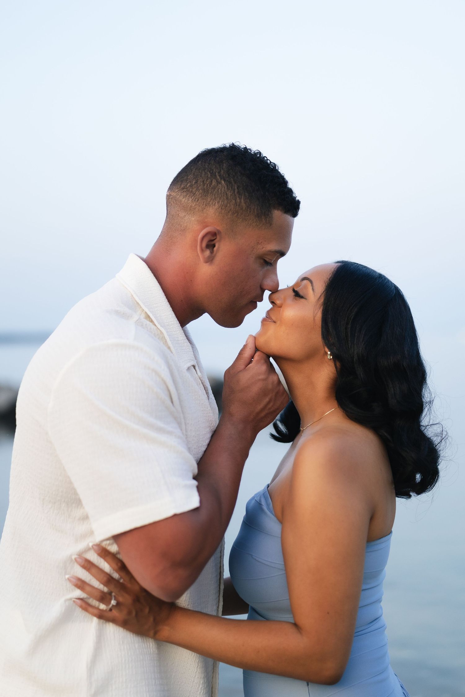 Romantic couple sharing a tender kiss near the water at dusk, man in white shirt gently holding womans face.