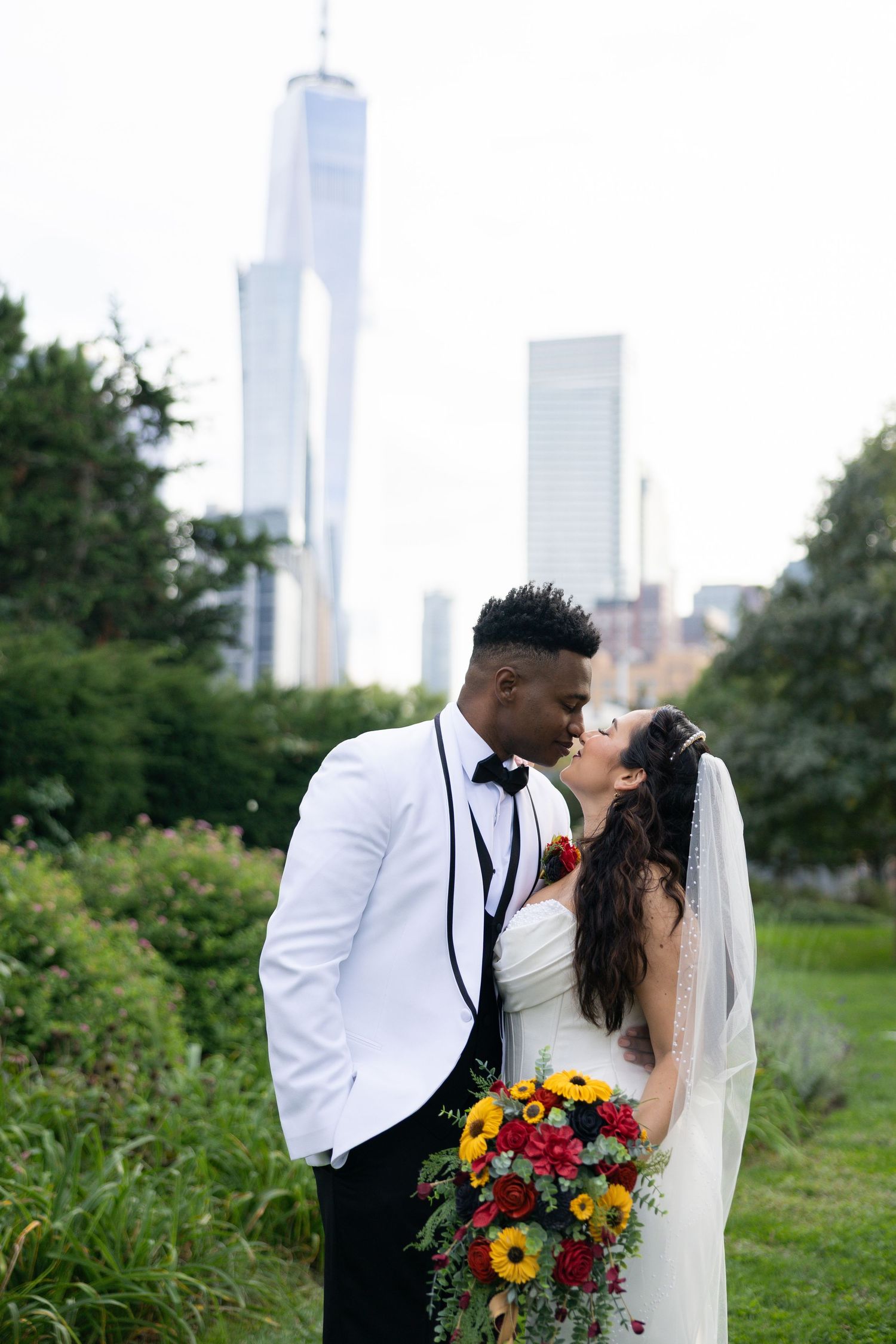 A newlywed couple shares a kiss in a garden with the NYC skyline and One World Trade Center in the background.