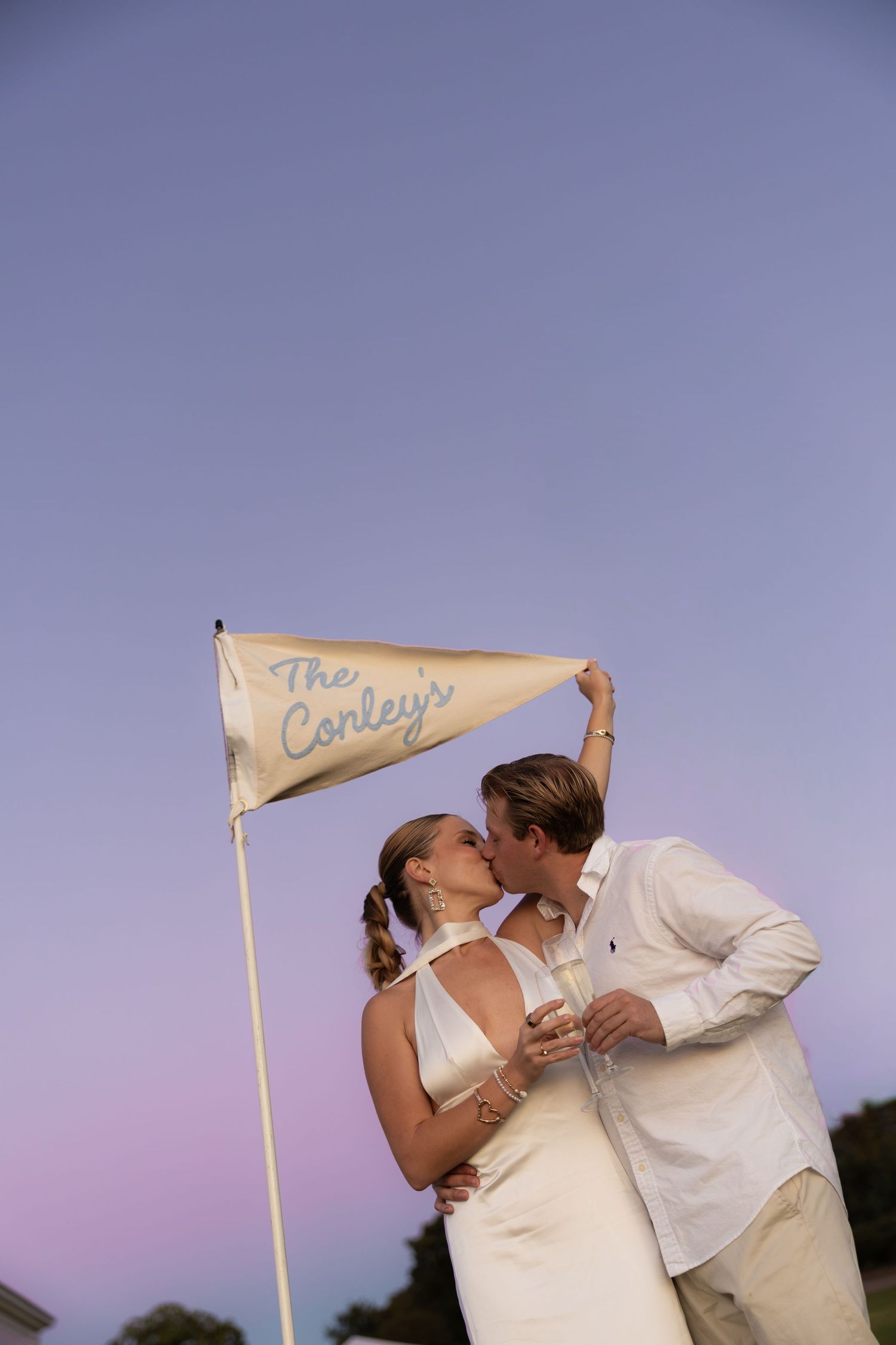 Newlywed couple kissing under a pennant flag reading The Conleys at dusk with a purple sky backdrop.