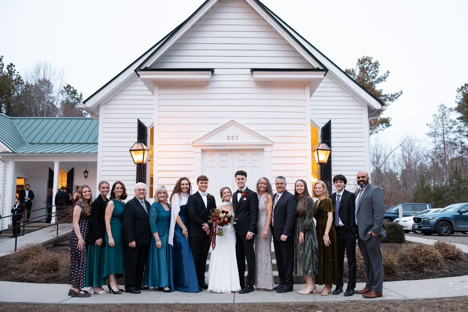 Large wedding party posing outside a white chapel venue with lanterns at dusk.