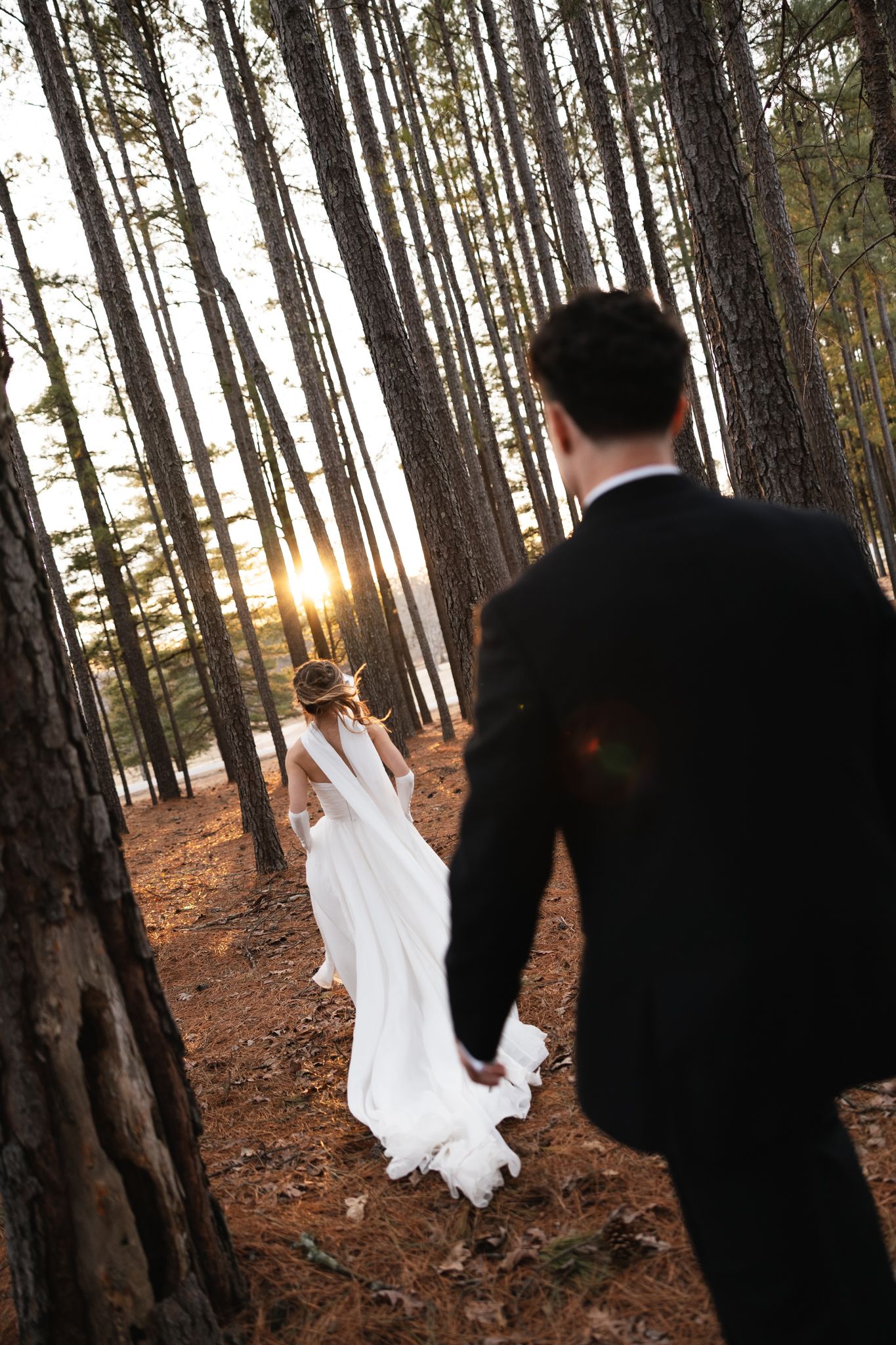 Bride and groom walk hand in hand through a pine forest at golden sunset on their wedding day.