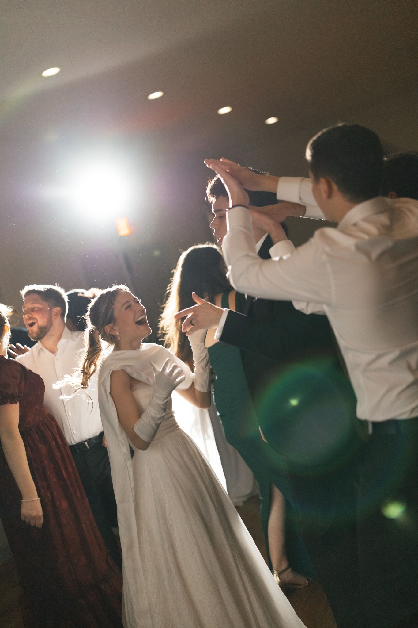 Bride in white gown laughing and dancing with guests under bright reception lights.