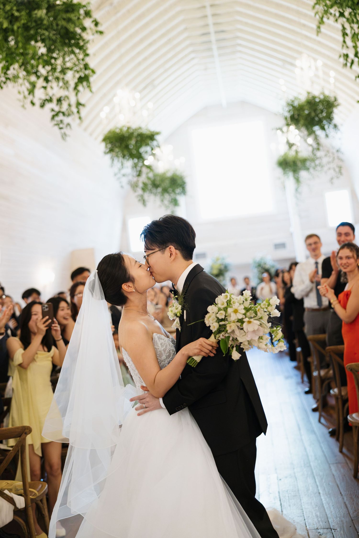 Bride and groom share first kiss at wedding ceremony aisle, guests applauding in decorated white chapel.