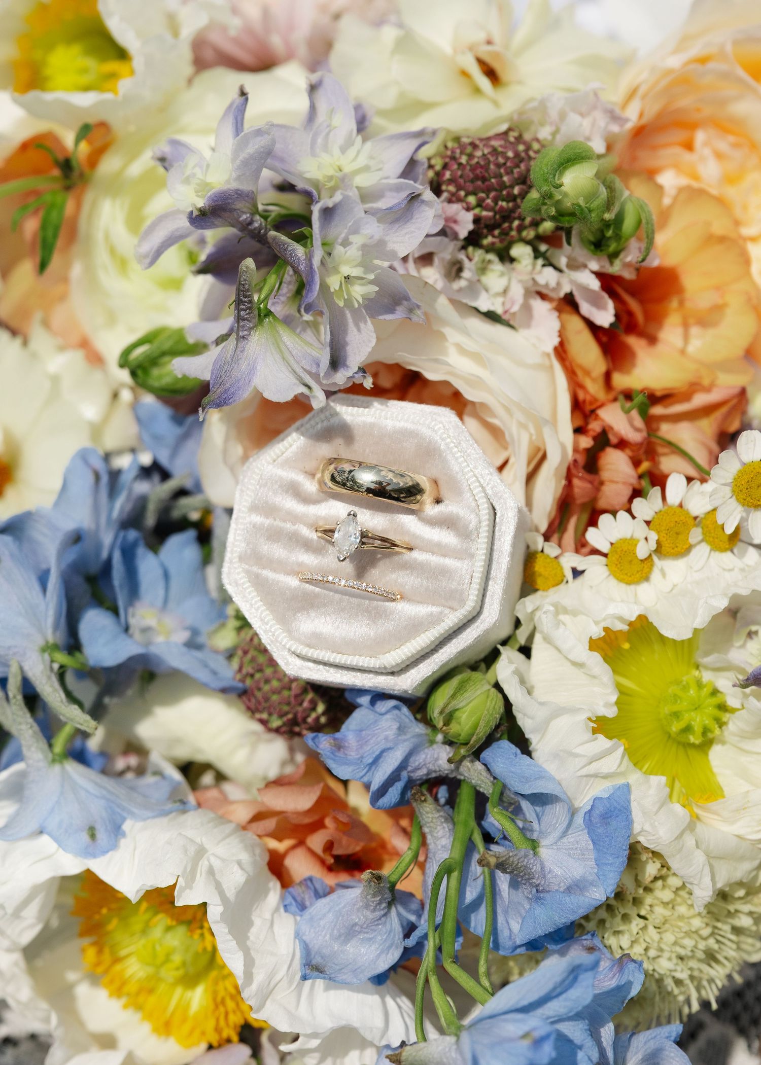 Wedding rings in a velvet box resting on a colorful bridal bouquet with blue, peach, and white flowers.