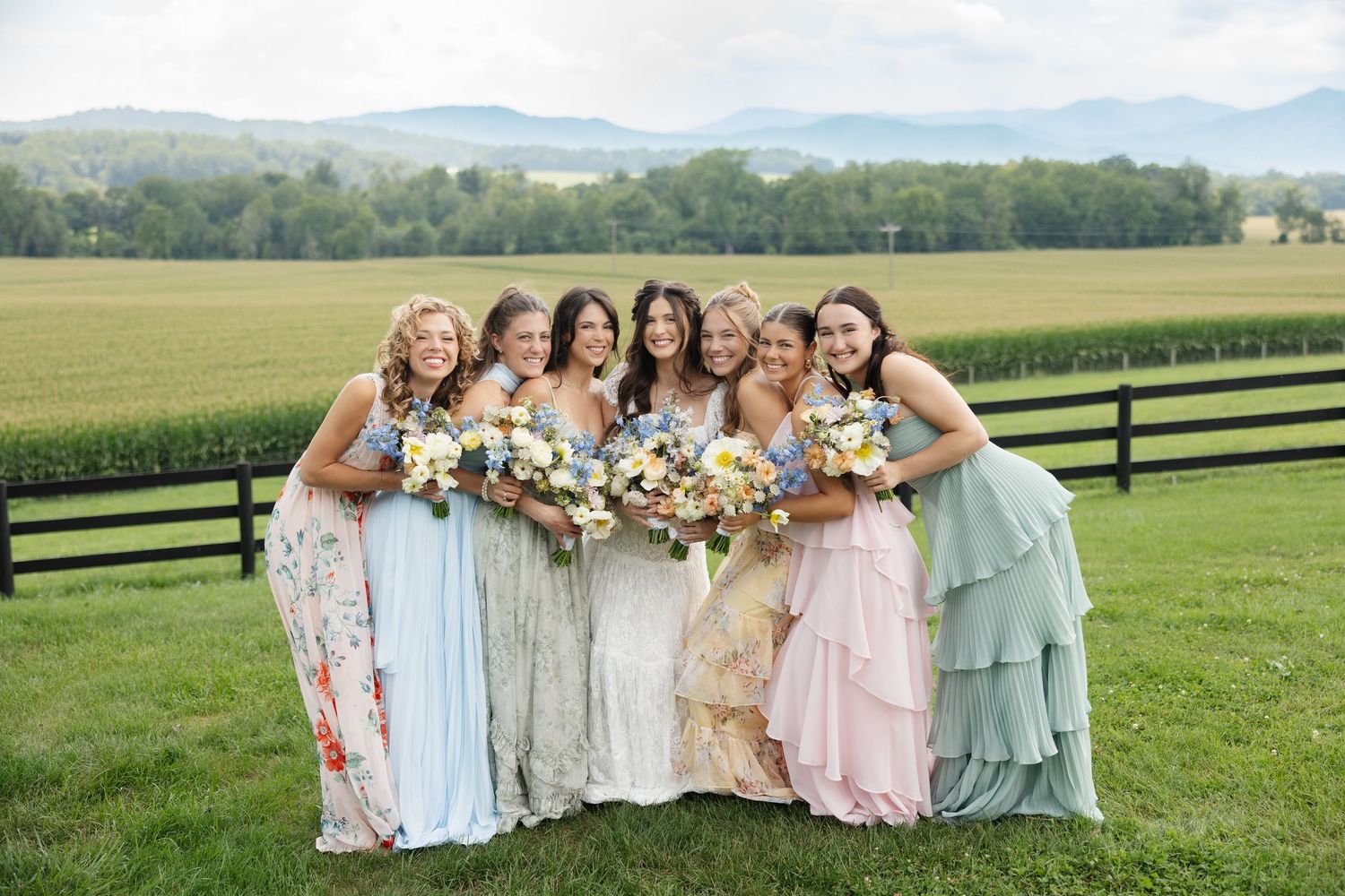 Bride and bridesmaids in colorful mismatched dresses holding bouquets on a green farm with mountain views.
