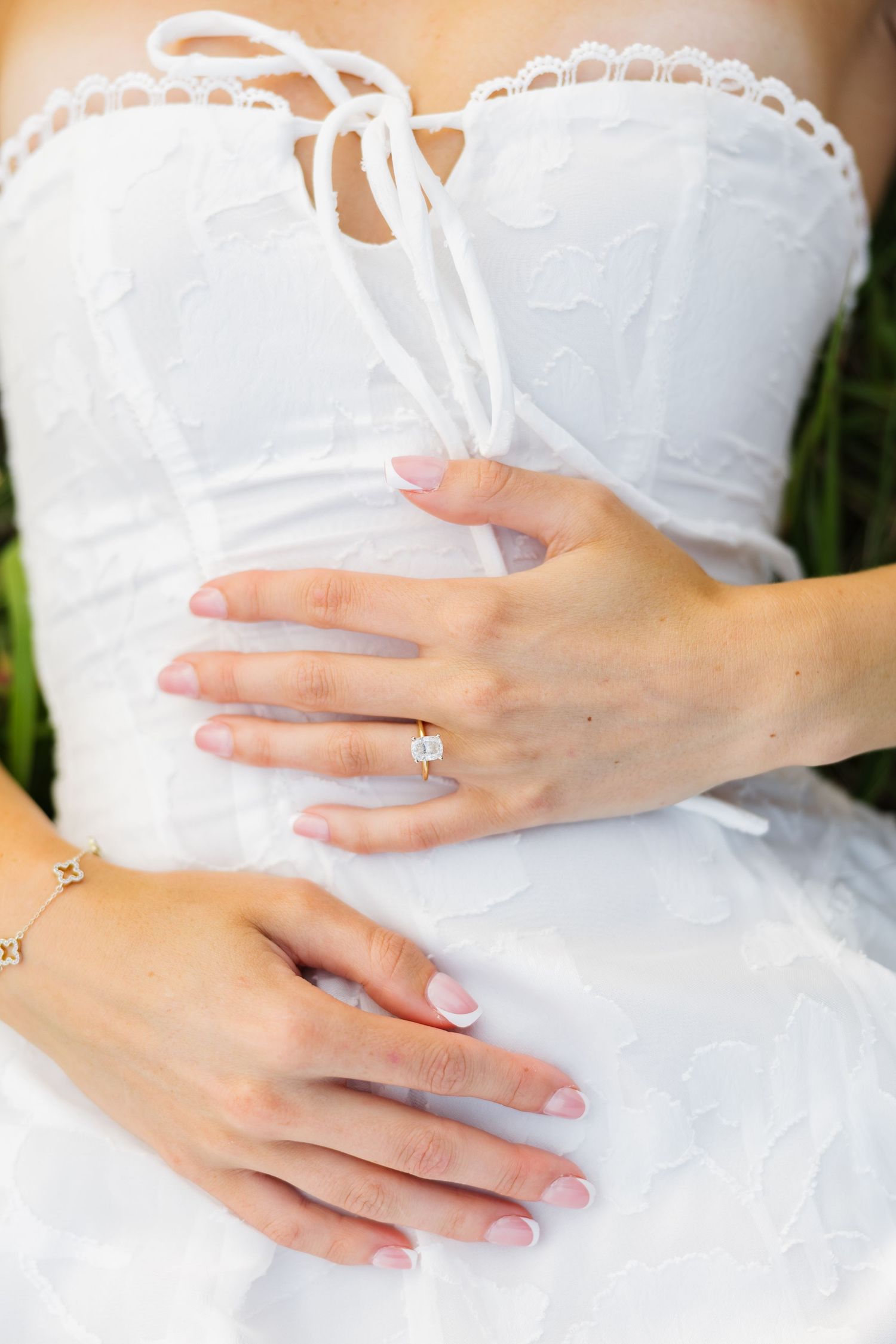 Bride in white lace dress with French manicure and diamond engagement ring resting hands on wedding gown.