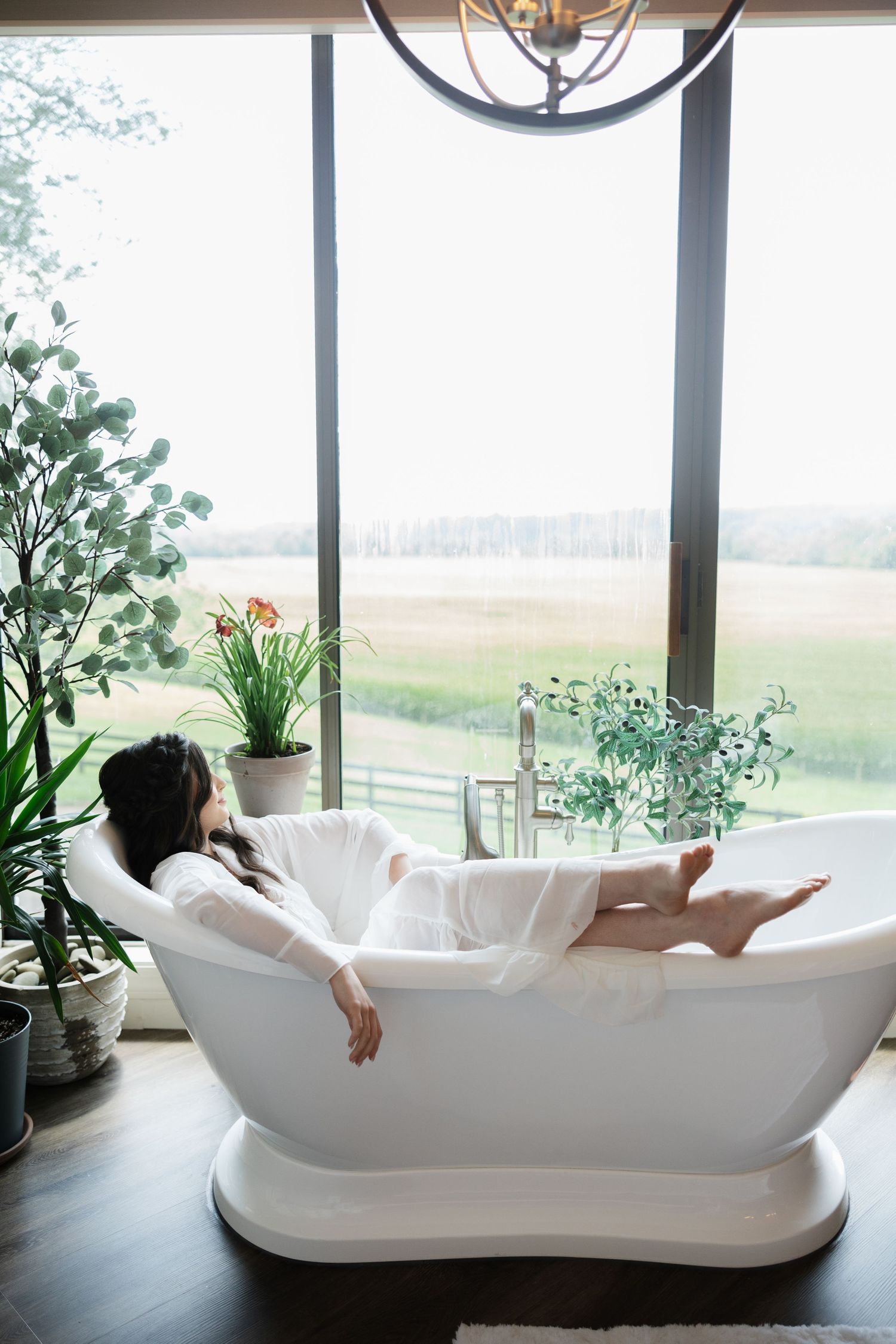 Woman in white robe relaxing in freestanding bathtub with scenic green field view through large windows.