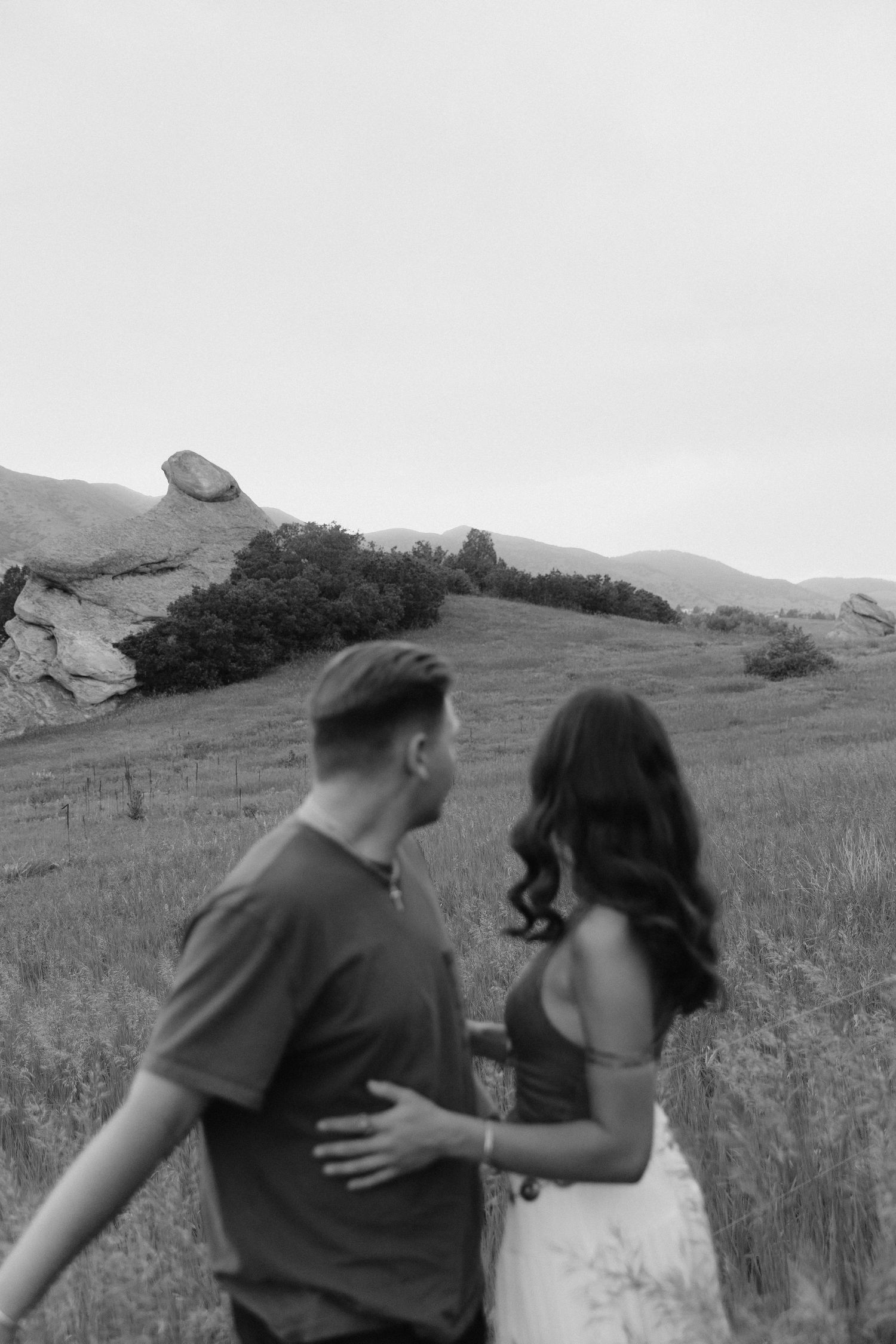 Black and white photo of a couple walking together through a scenic field with rocky mountain backdrop.