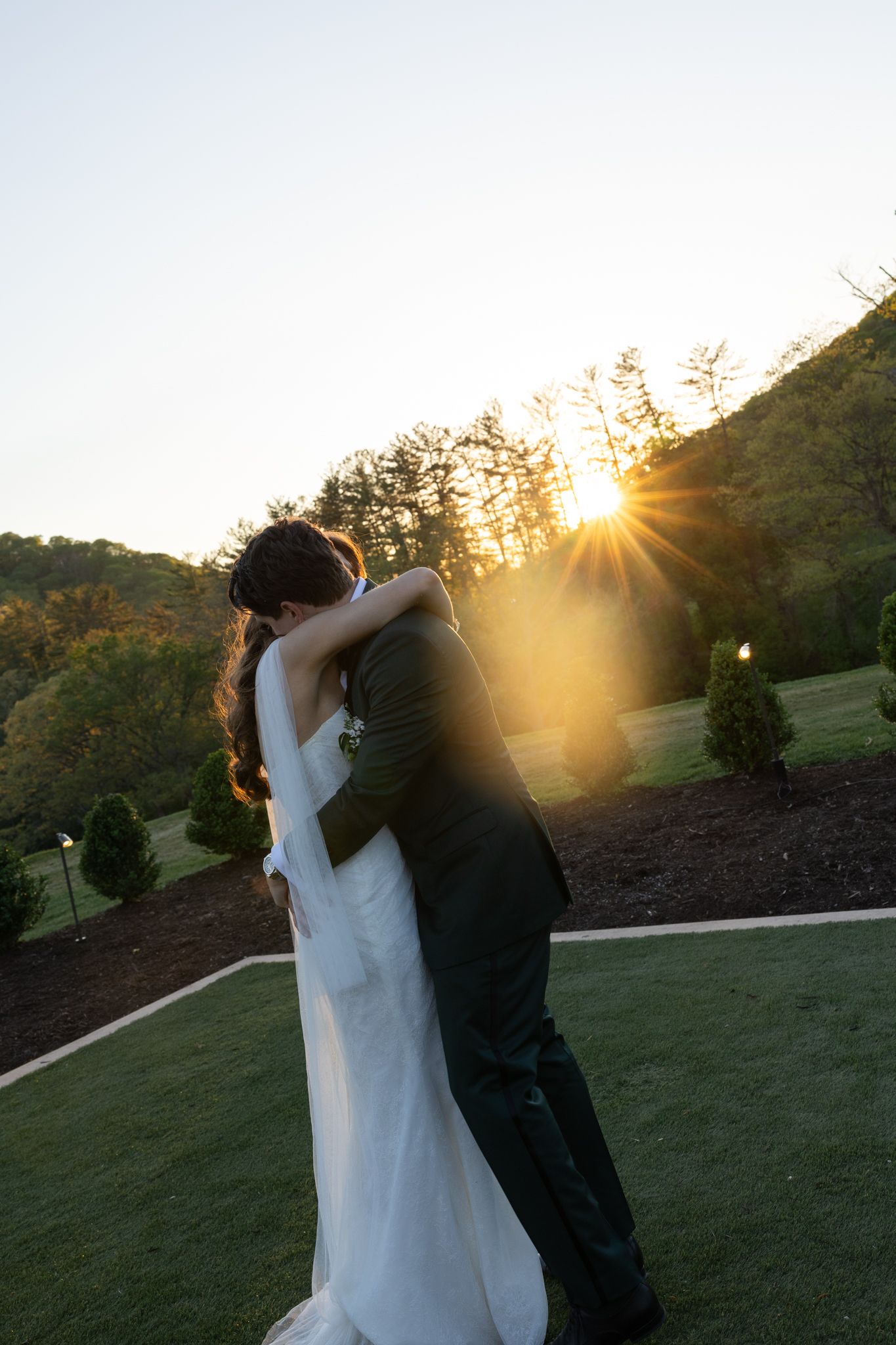 Bride and groom embracing at sunset with golden sunburst light on a lush green lawn.