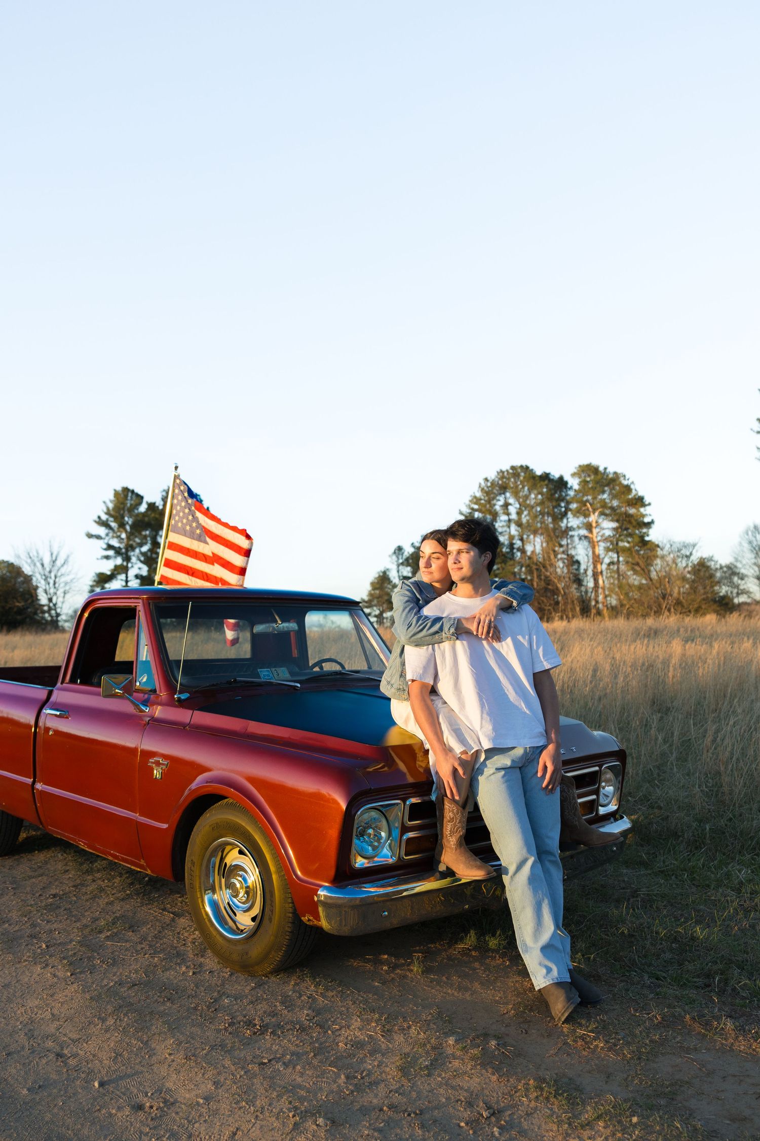 Couple embracing beside a classic red pickup truck with an American flag in an open field.