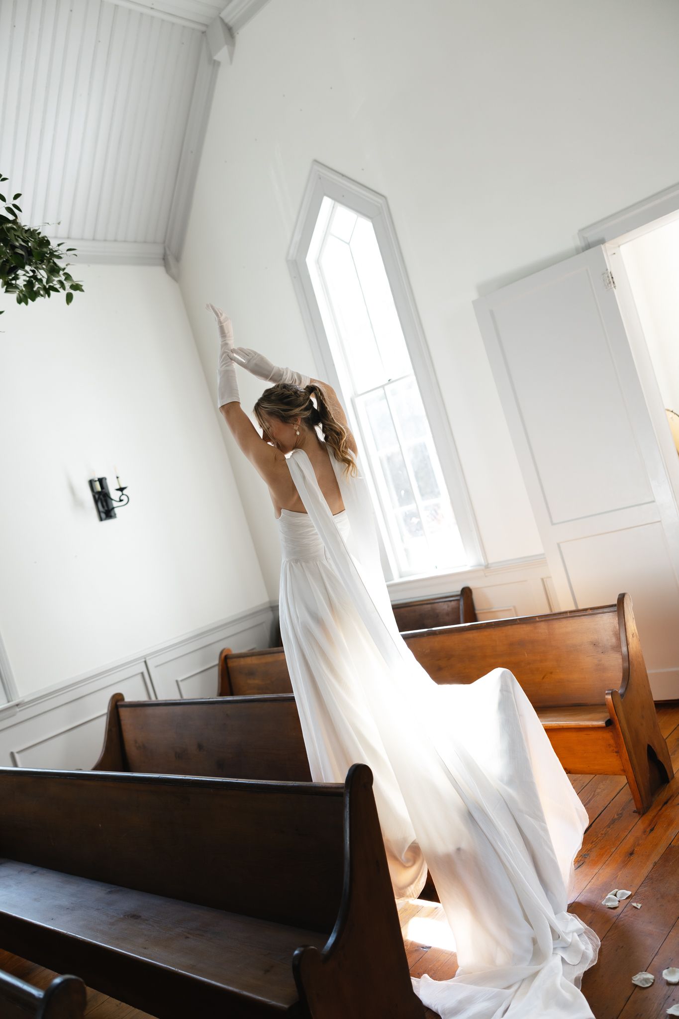Bride in white wedding gown twirling in a sunlit chapel aisle between wooden pews.