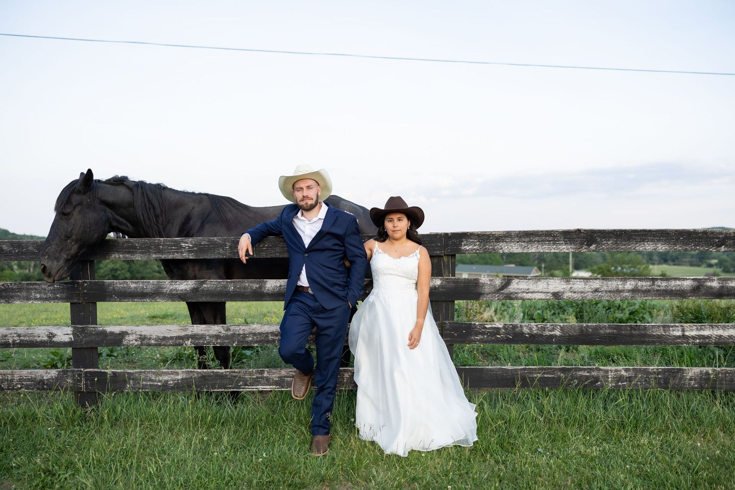 Bride and groom in cowboy hats pose by a rustic wooden fence with a black horse on a green farm.
