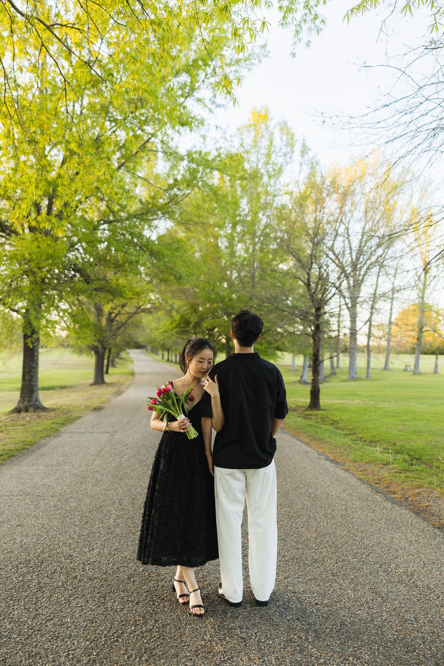 A couple walks down a tree-lined path, the woman in a black dress smelling a bouquet of red tulips.