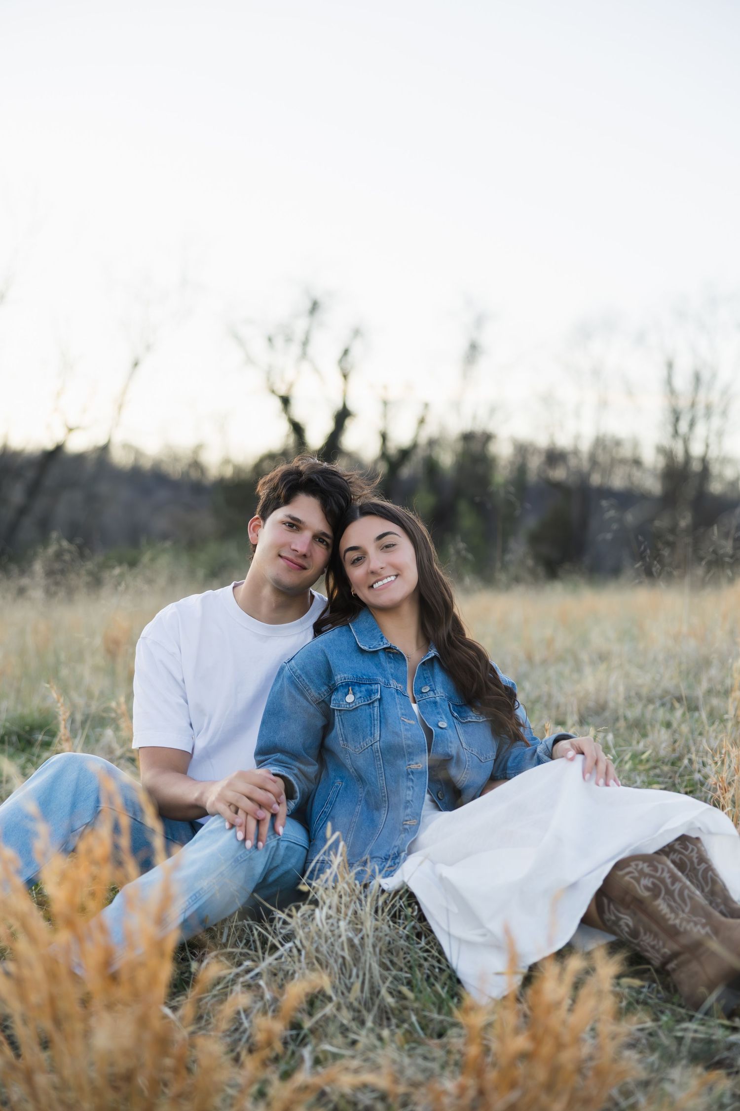 A smiling couple sitting together in a golden grass field at sunset, the woman in a denim jacket and white skirt.