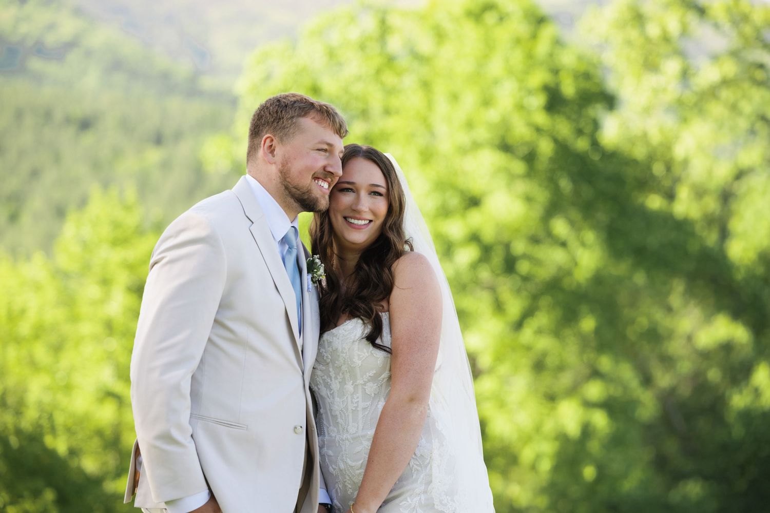 Happy bride and groom pose together outdoors with lush green mountain landscape behind them on their wedding day.