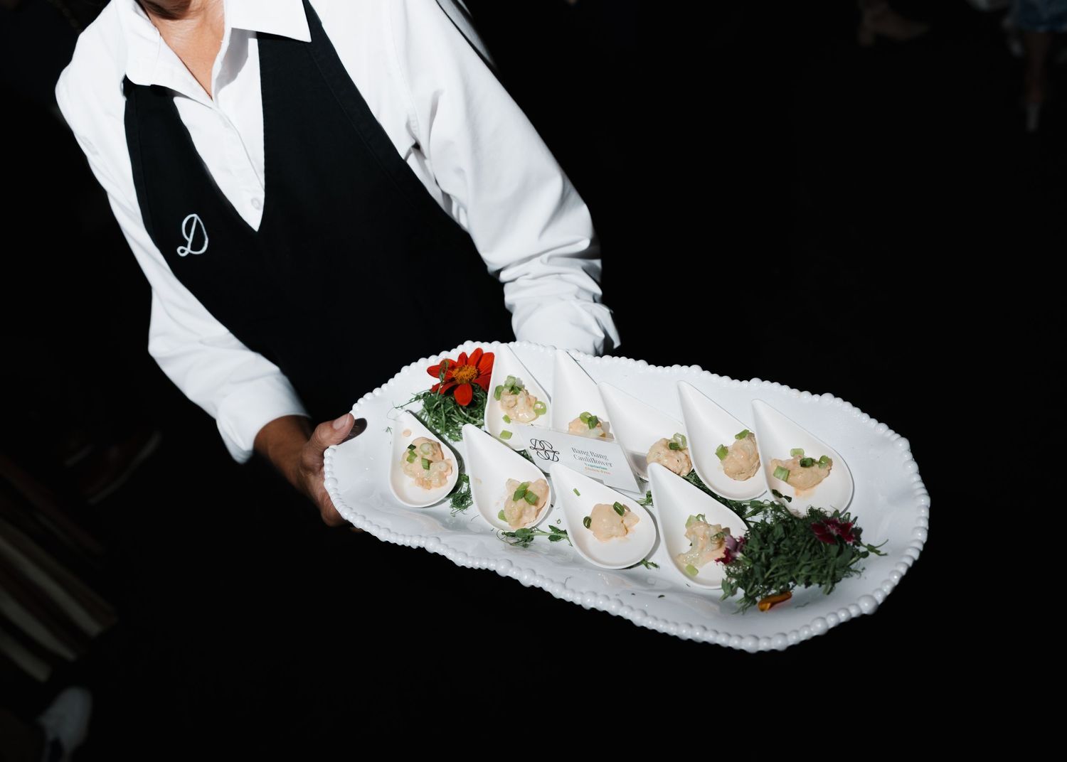 Waiter in black vest holding a white platter with elegant appetizers and red floral garnish at a catered event.