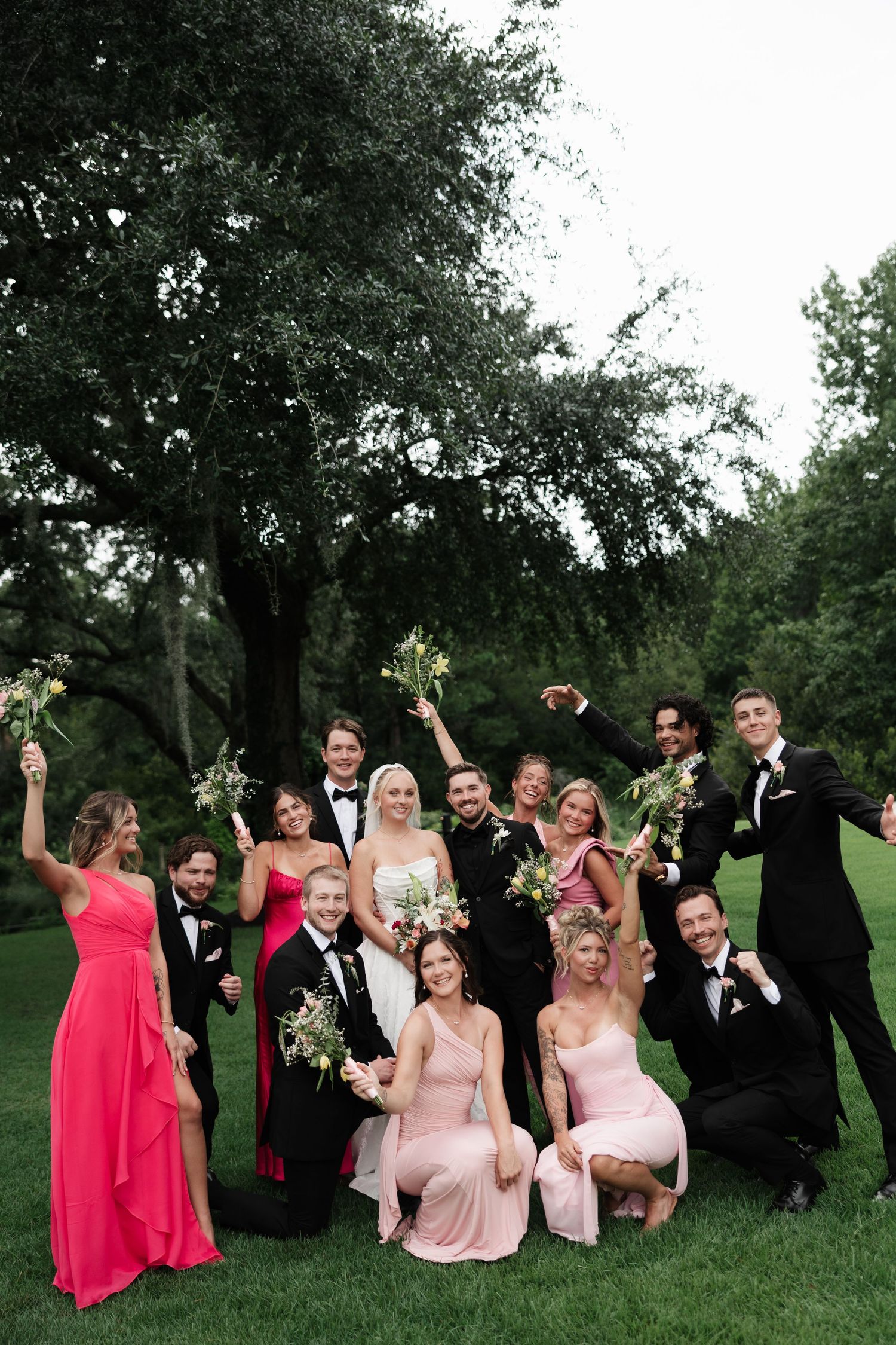Wedding party posing outdoors on green lawn, bridesmaids in pink and red gowns, groomsmen in black tuxedos, bride in white gown.