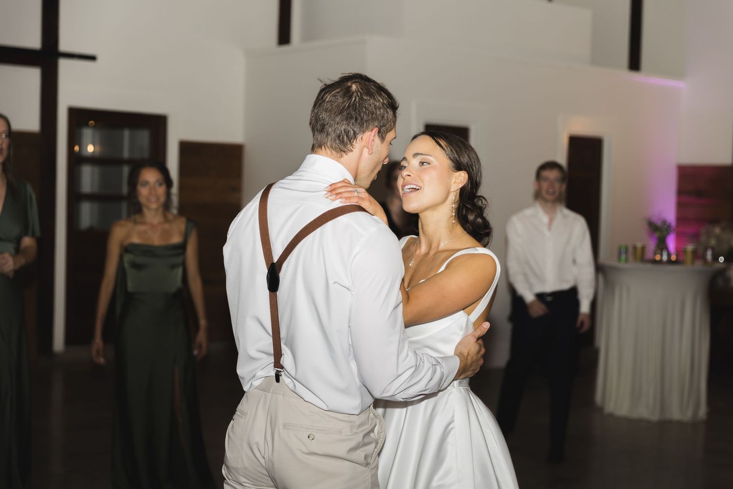 Bride and groom share their first dance in a warmly lit reception hall with guests watching nearby.