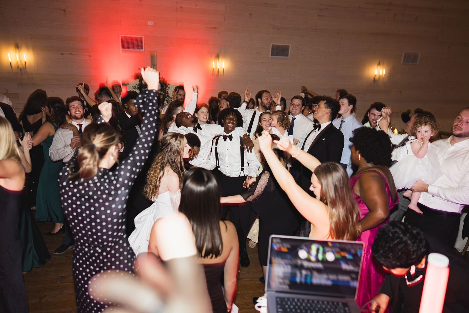 Guests and wedding party dance together on a crowded dance floor with red ambient lighting.