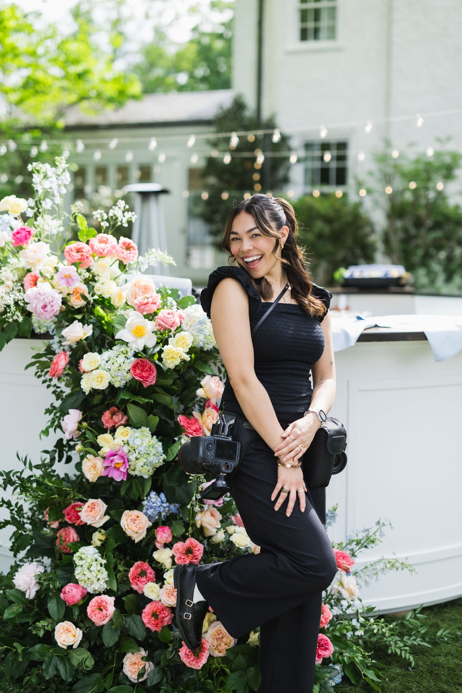 Woman in black outfit smiling beside colorful floral arrangement at outdoor garden event with string lights.