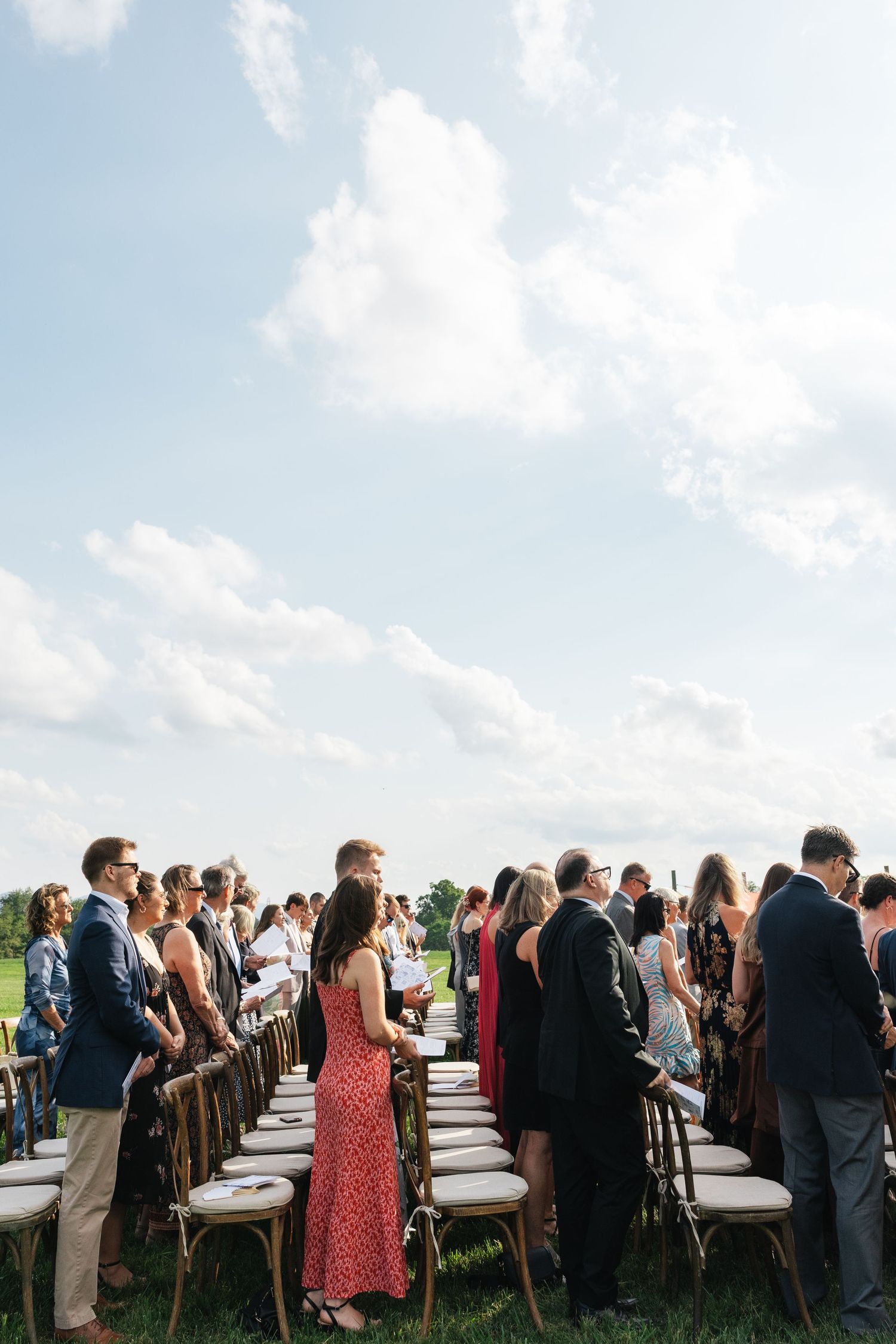 Guests standing at an outdoor wedding ceremony on a sunny day with rows of wooden chairs on green grass.