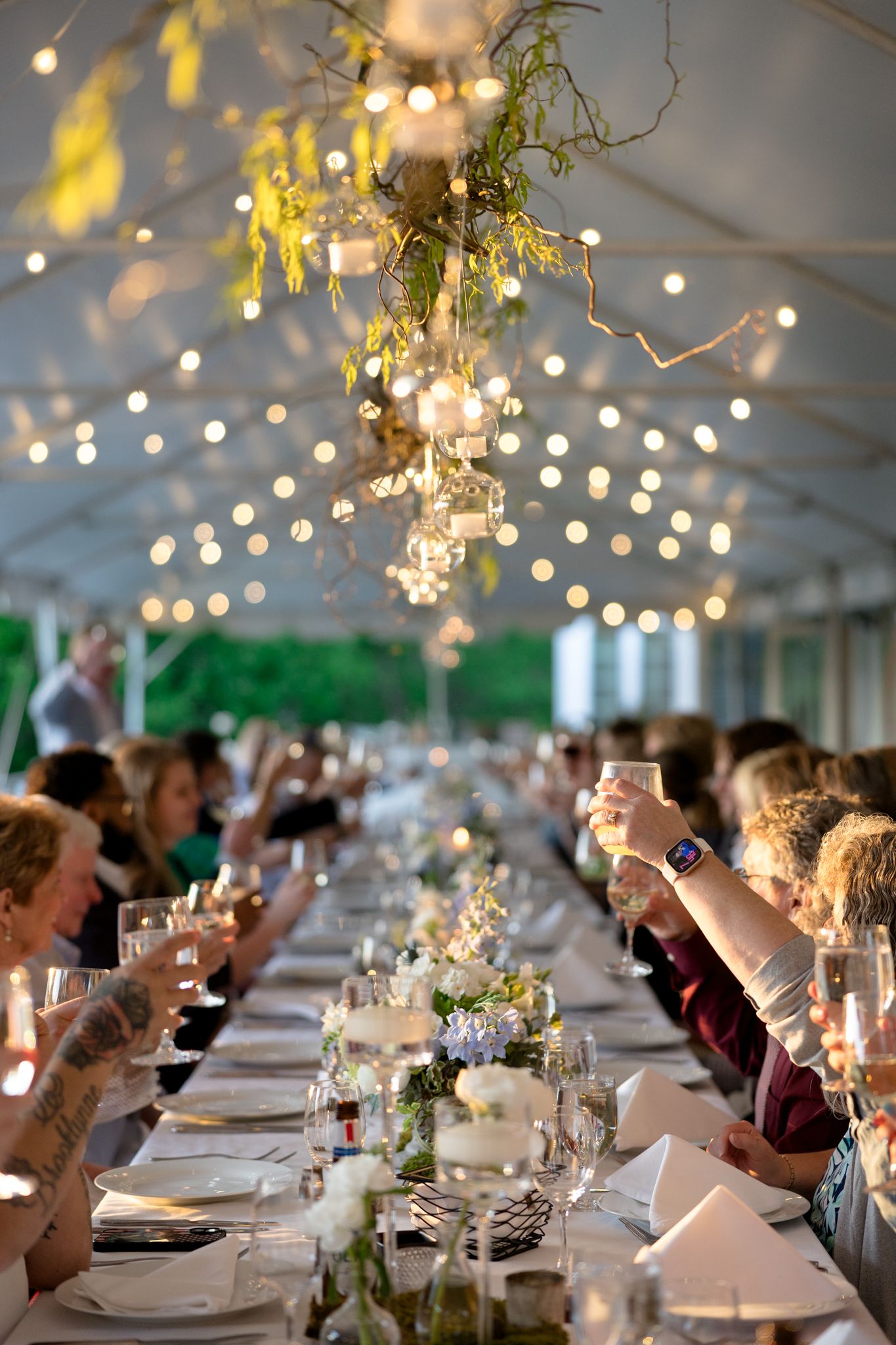 Rehearsal dinner guests raise their glasses in a toast under a white tent adorned with string lights and floral arrangements.