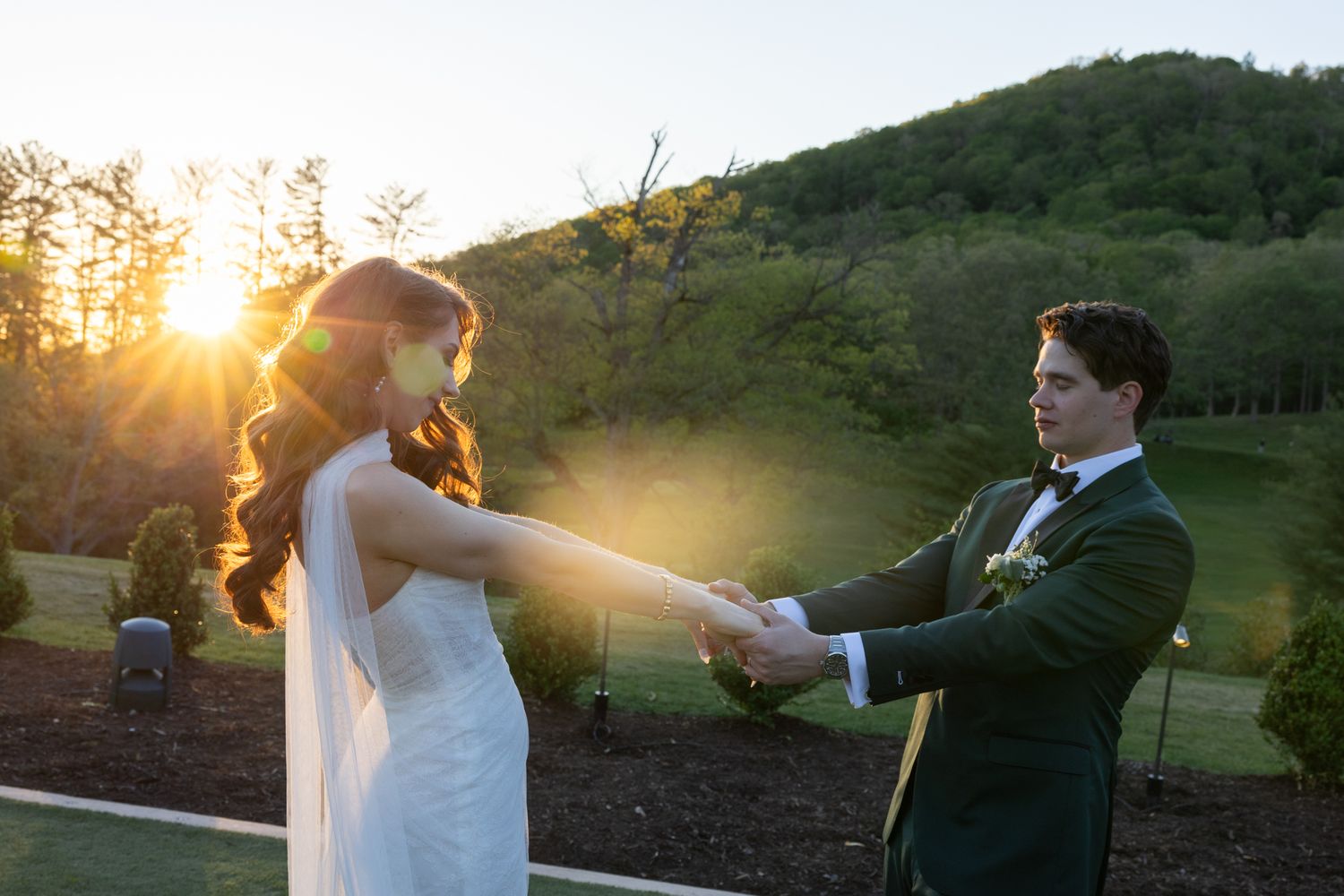 Bride and groom holding hands at golden hour sunset with lush green hills in background at outdoor wedding ceremony.
