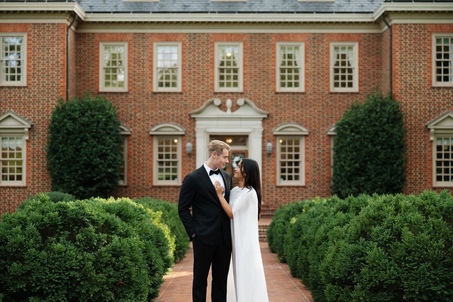 Couple in wedding attire embracing before a grand brick colonial building surrounded by manicured hedges.