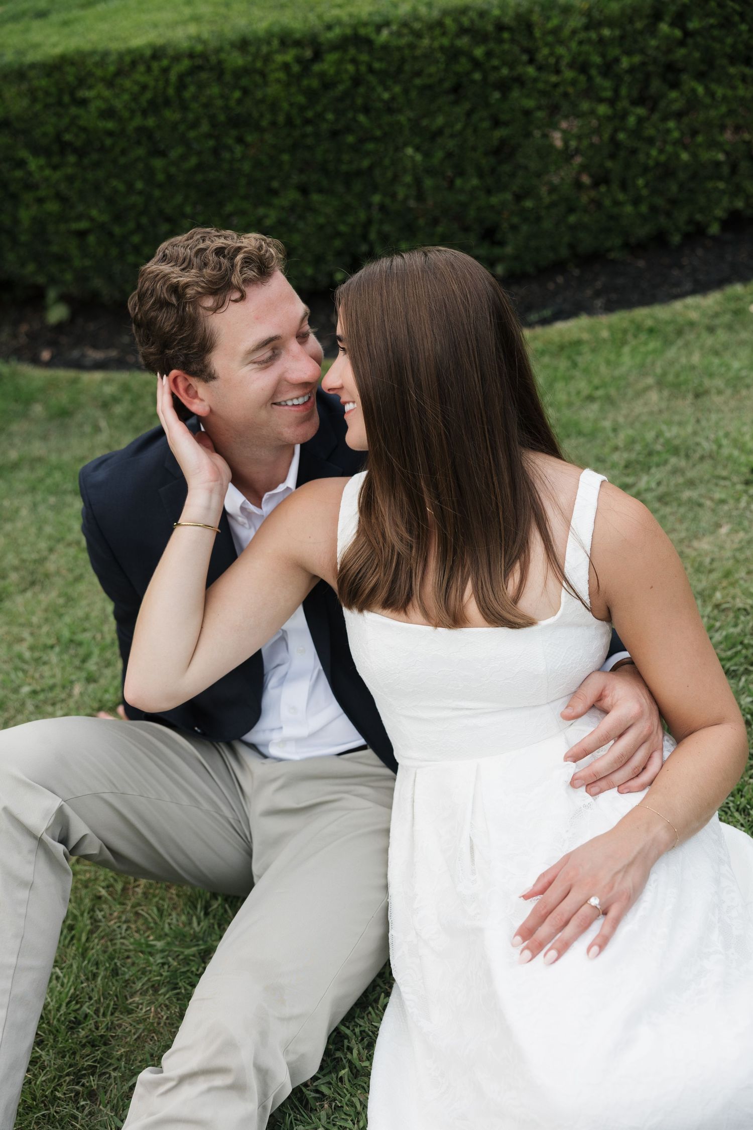 Couple sitting on grass, woman in white dress and man in navy blazer share a romantic moment outdoors.