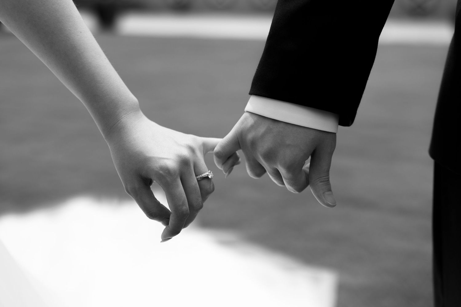 Black and white photo of a bride and groom holding hands, showing wedding rings, in a romantic outdoor setting.