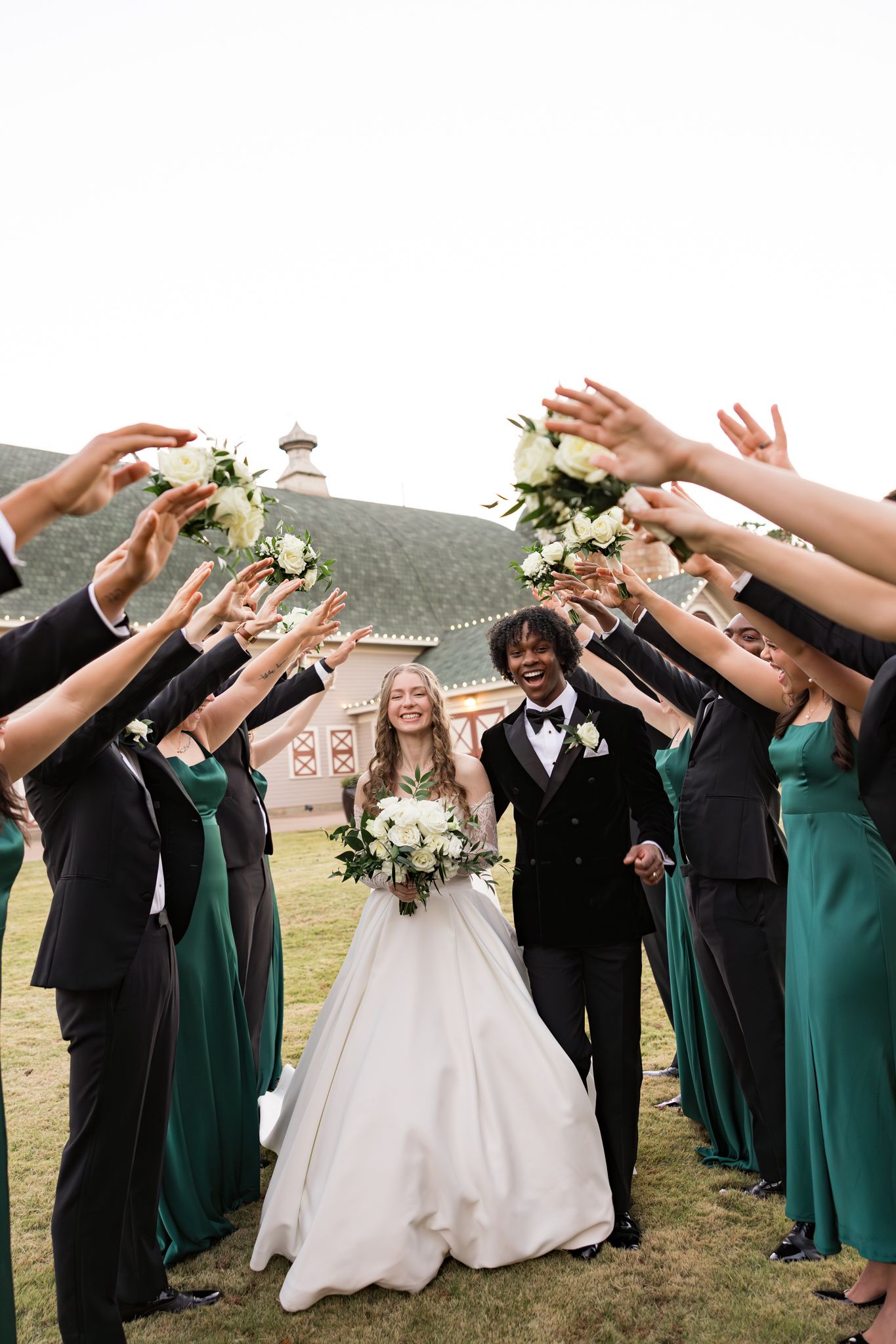 Bride and groom walk through arch of raised bouquets held by wedding party outside barn.