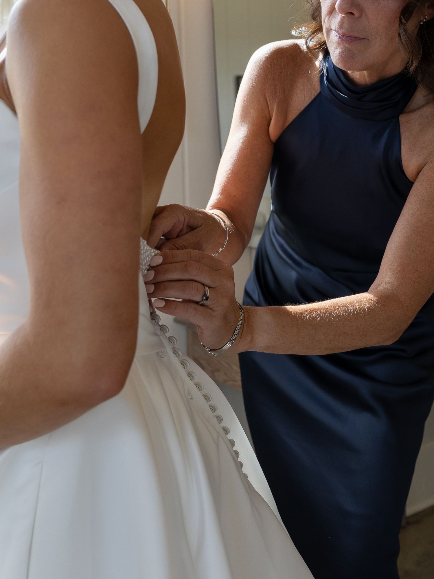 Woman in navy dress carefully buttoning the back of a bride's white wedding gown.