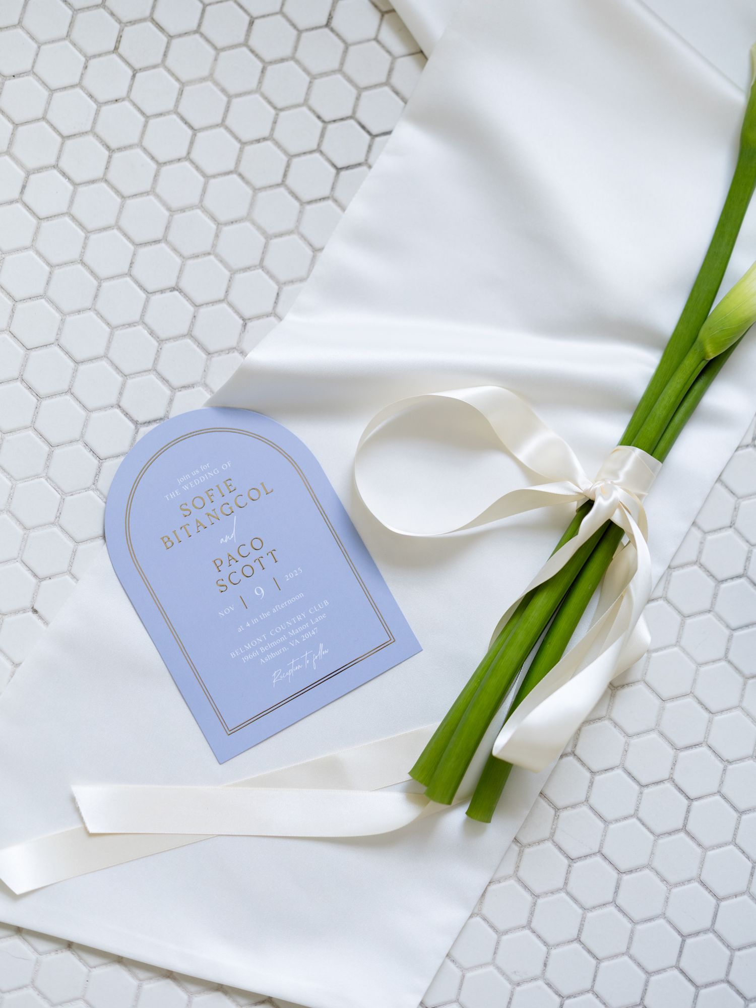 Elegant blue and gold arch wedding invitation next to calla lily stems tied with white satin ribbon.