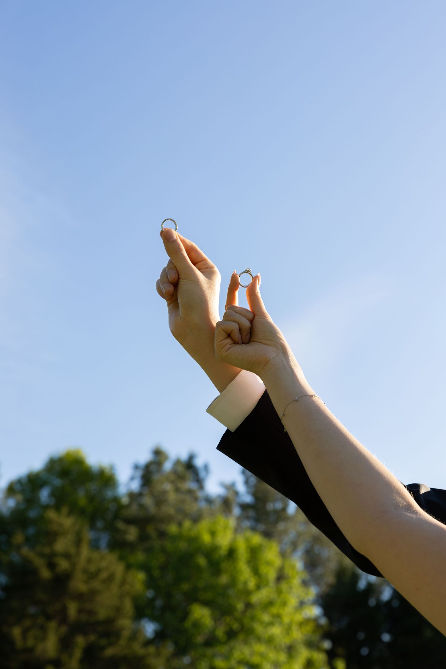 Two hands hold wedding rings up against a clear blue sky with trees in background.