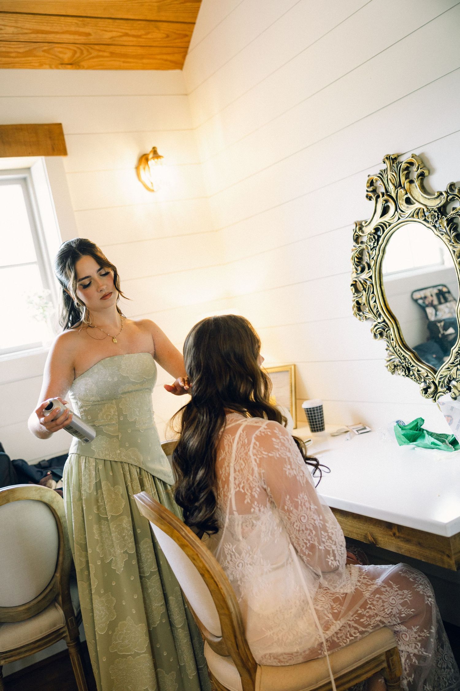 Bridesmaid in sage green dress styling bride's hair before wedding.