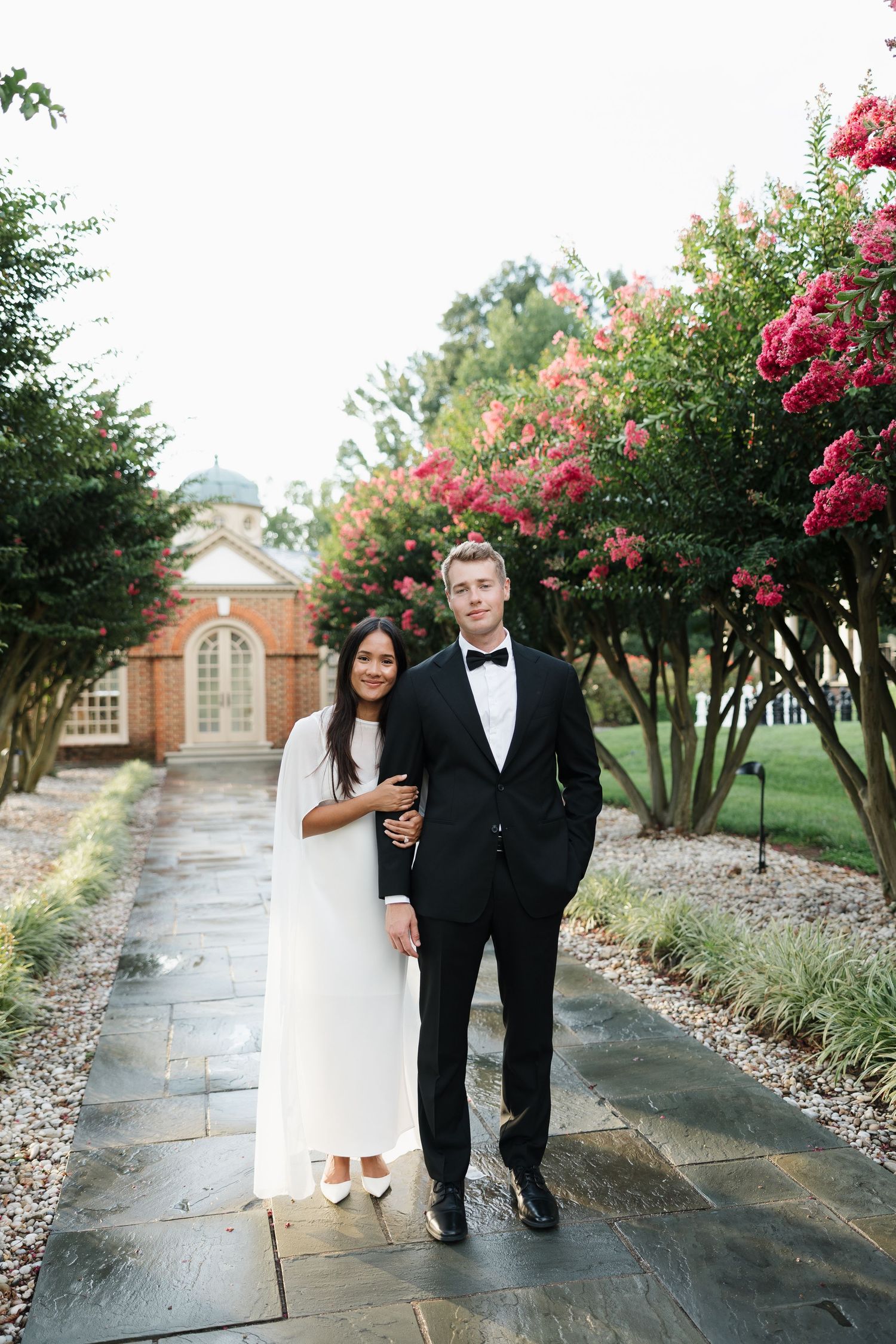 Couple in wedding attire posing on stone path with blooming pink crepe myrtle trees and brick building backdrop.